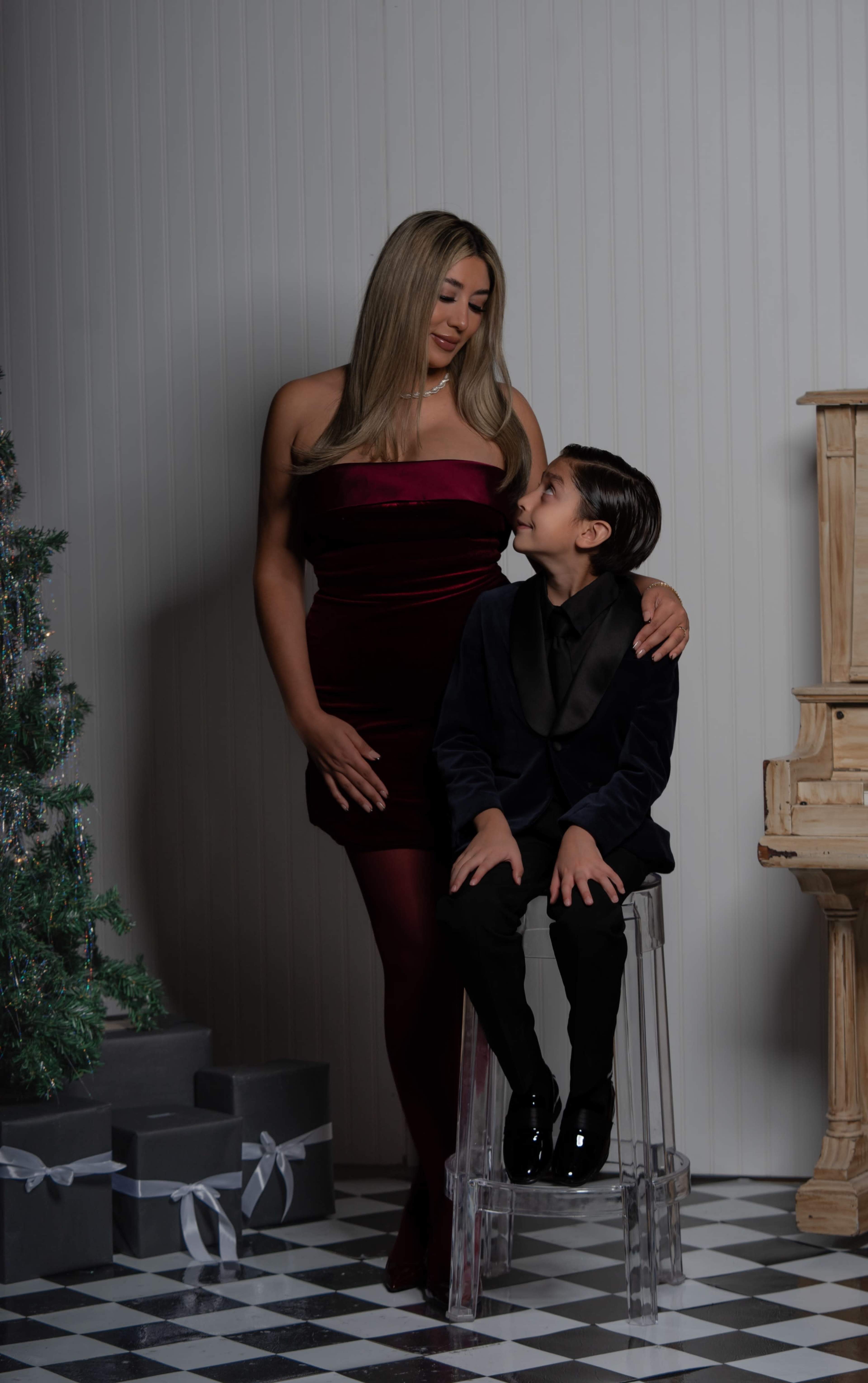 A woman in a burgundy dress stands beside a boy in a black suit, both looking at each other against a backdrop of a decorated Christmas tree and wrapped gifts.