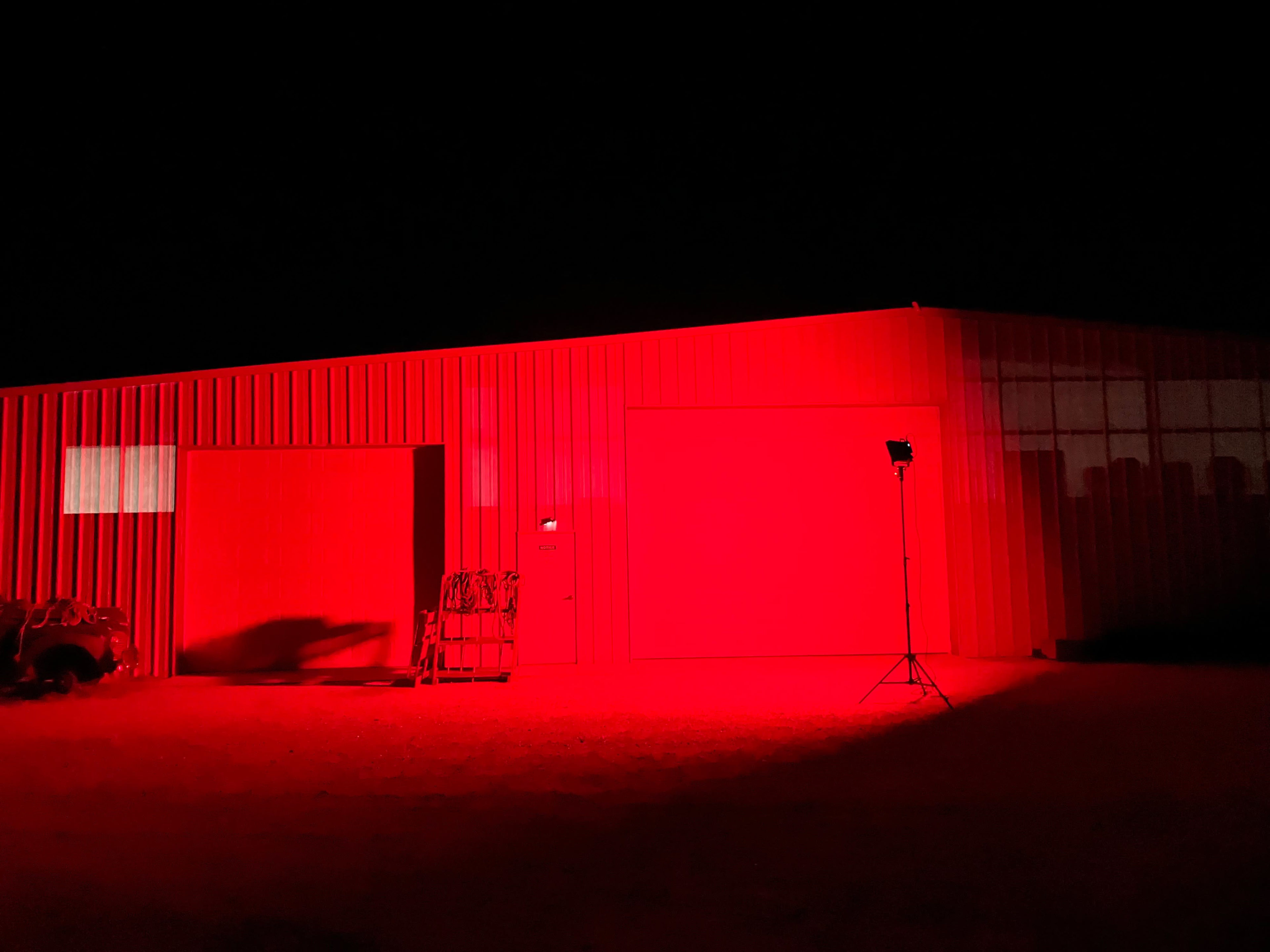 A metal building is illuminated with red light at night, highlighting two large doors and a nearby tripod-mounted light.