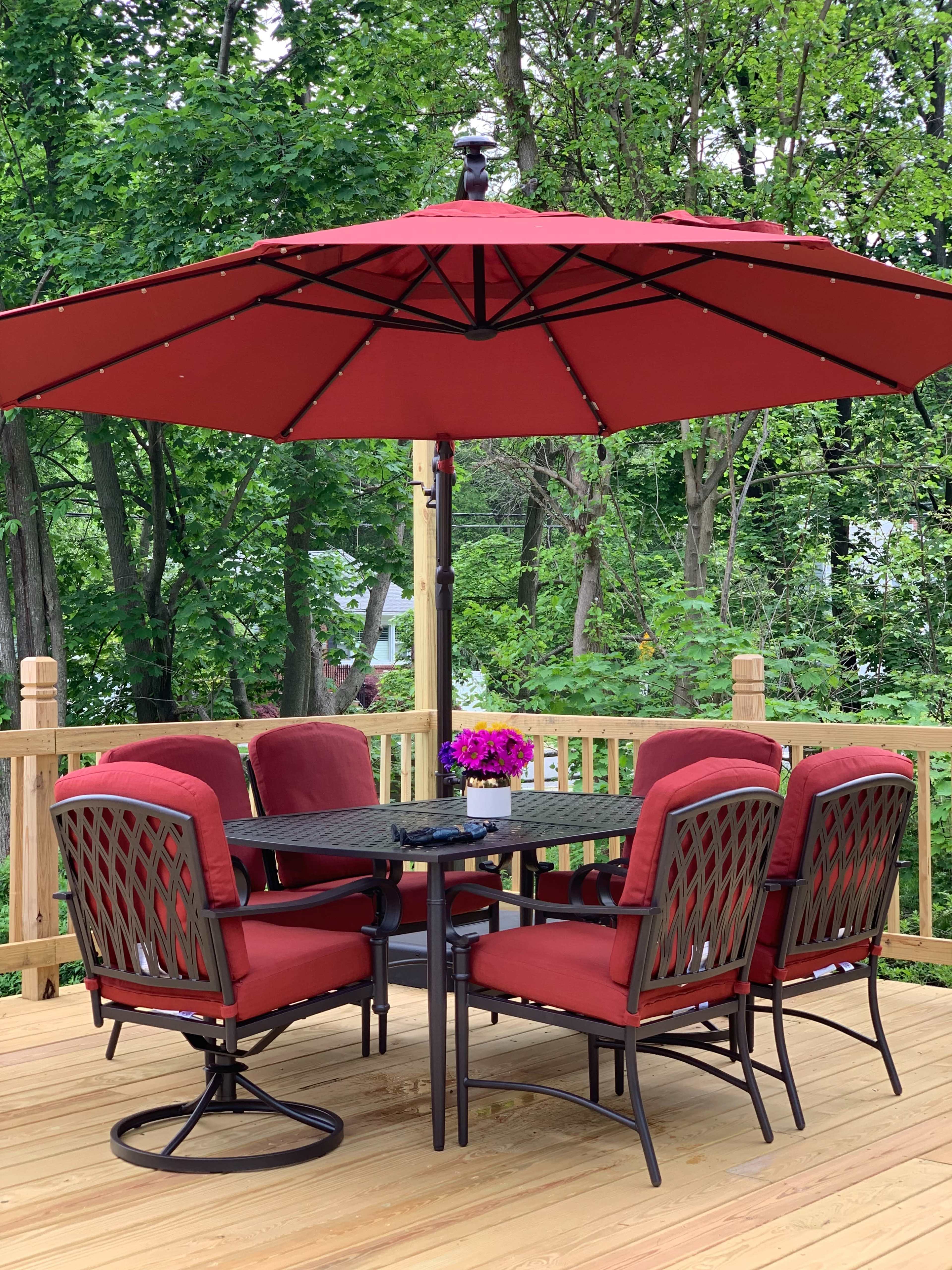 A red patio umbrella shades a set of six cushioned chairs around a black metal table on a wooden deck surrounded by greenery.