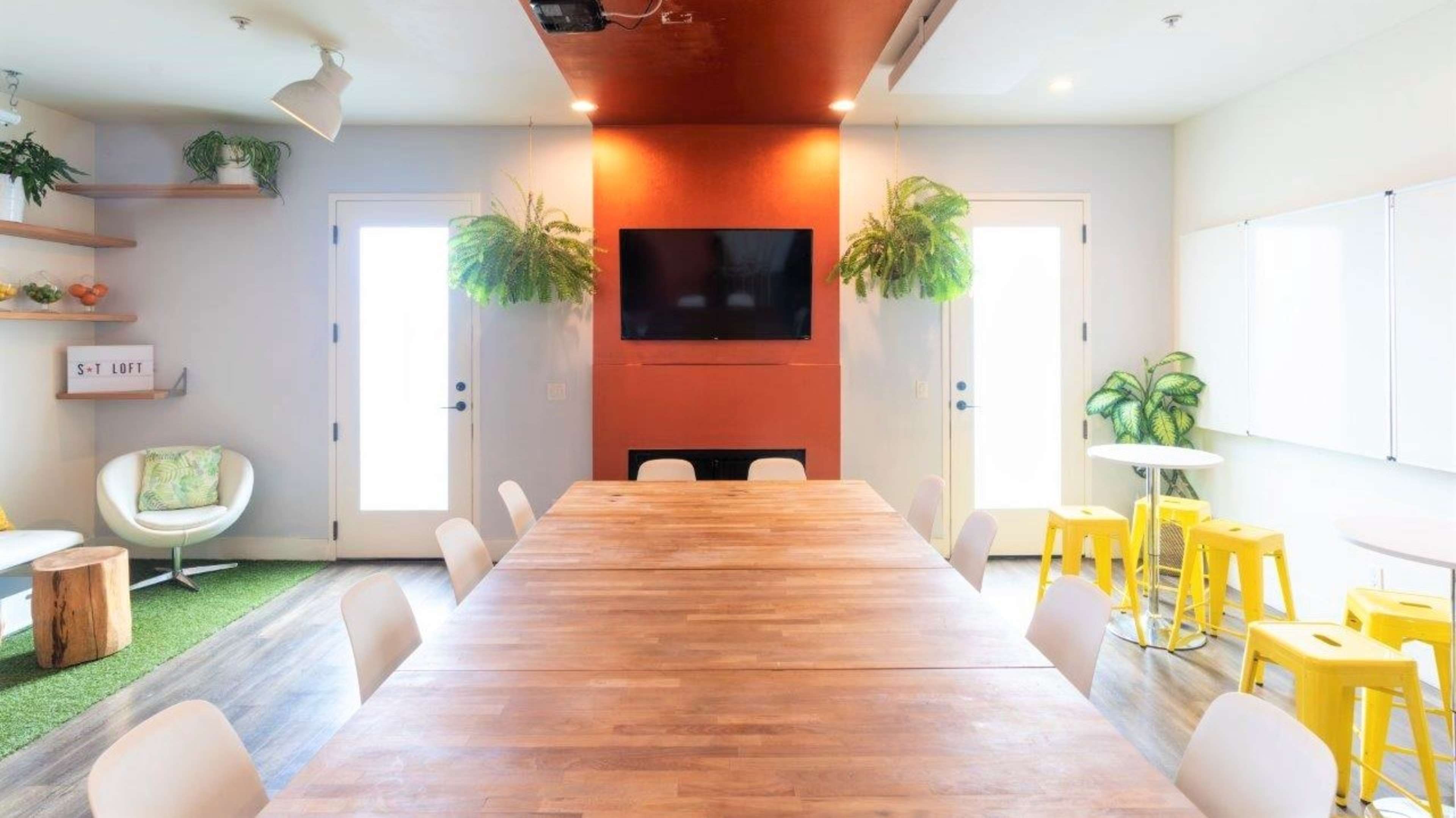 A large wooden table is centered in a bright meeting room featuring potted plants, a flat-screen TV, and glass doors.