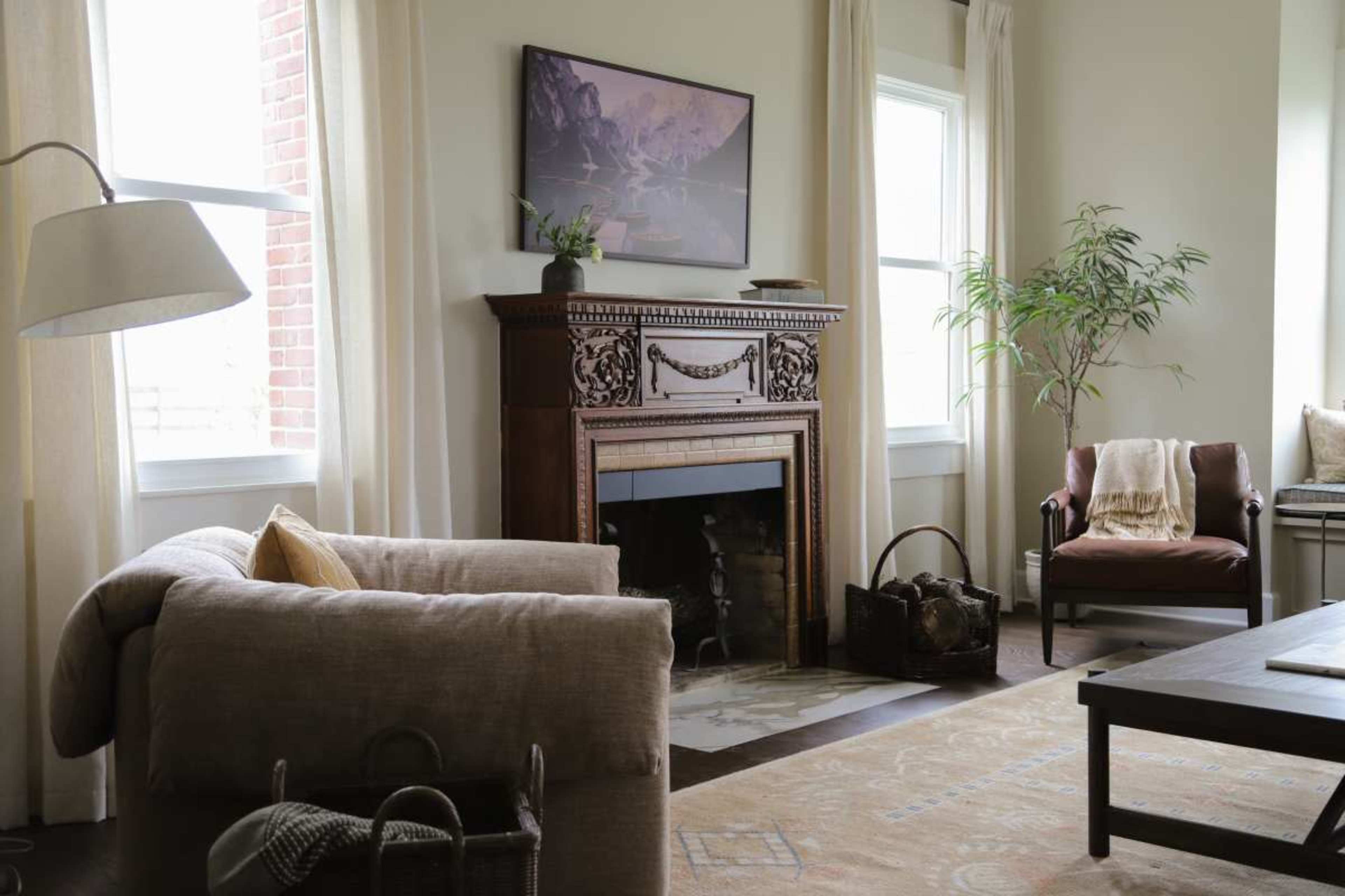 The living room features a fireplace with ornate detailing, a cozy sofa, a leather armchair, and large windows allowing natural light to enter.