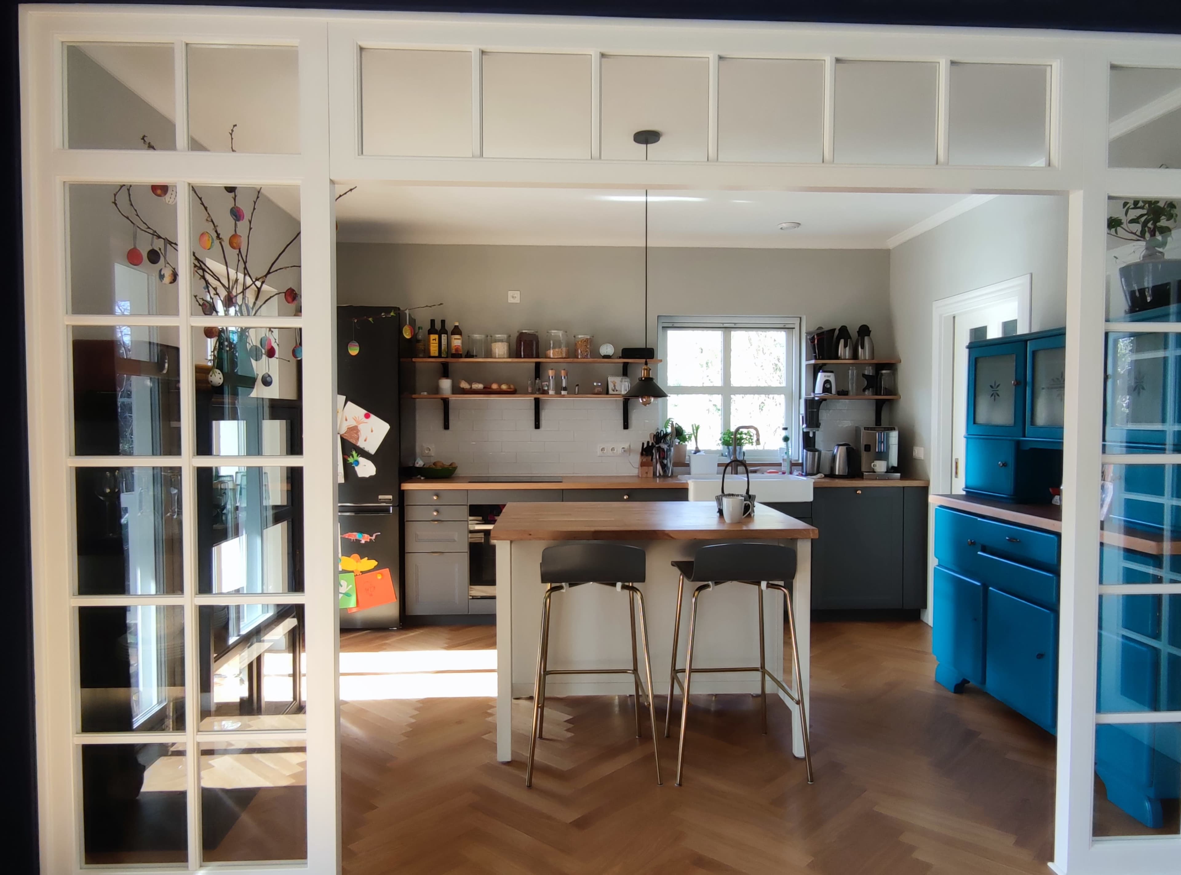 The image shows a modern kitchen with gray cabinets, a wooden countertop, and two stools, framed by a set of glass-panelled double doors.