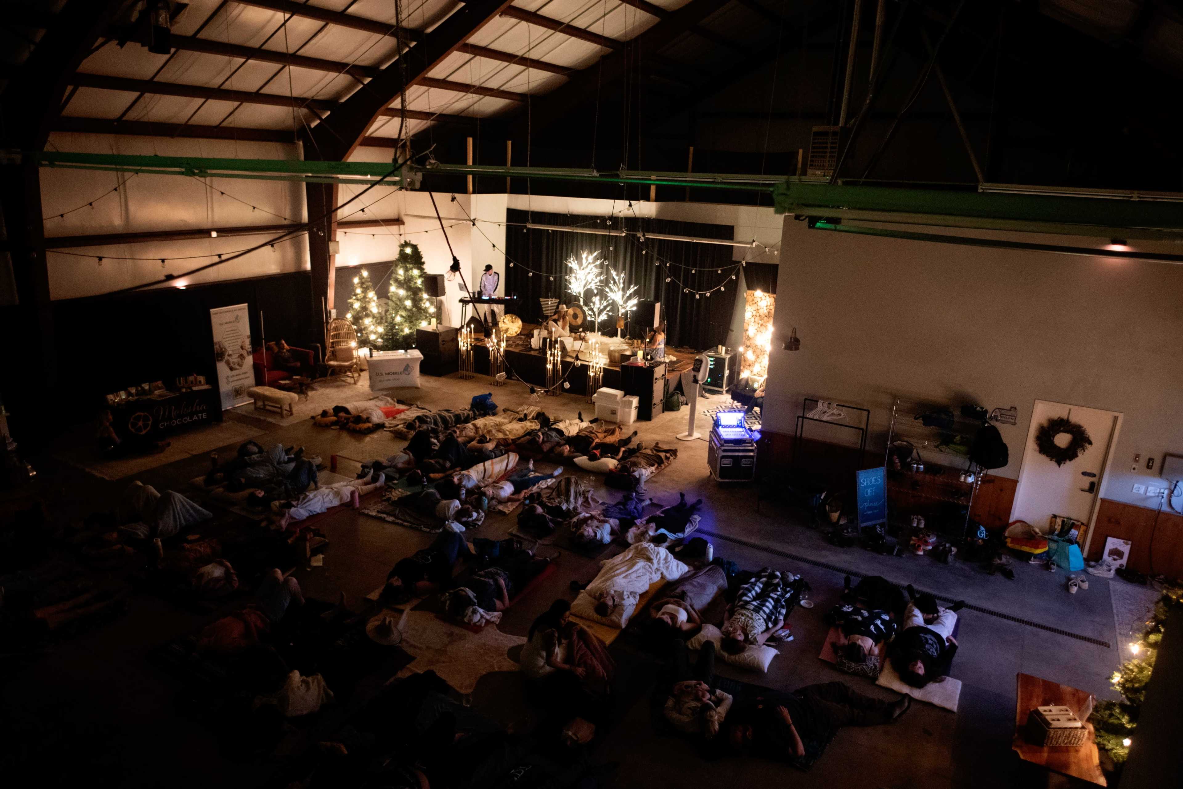 A large group of people lies on mats in a dimly lit warehouse, surrounded by holiday decorations and a stage with equipment in the background.