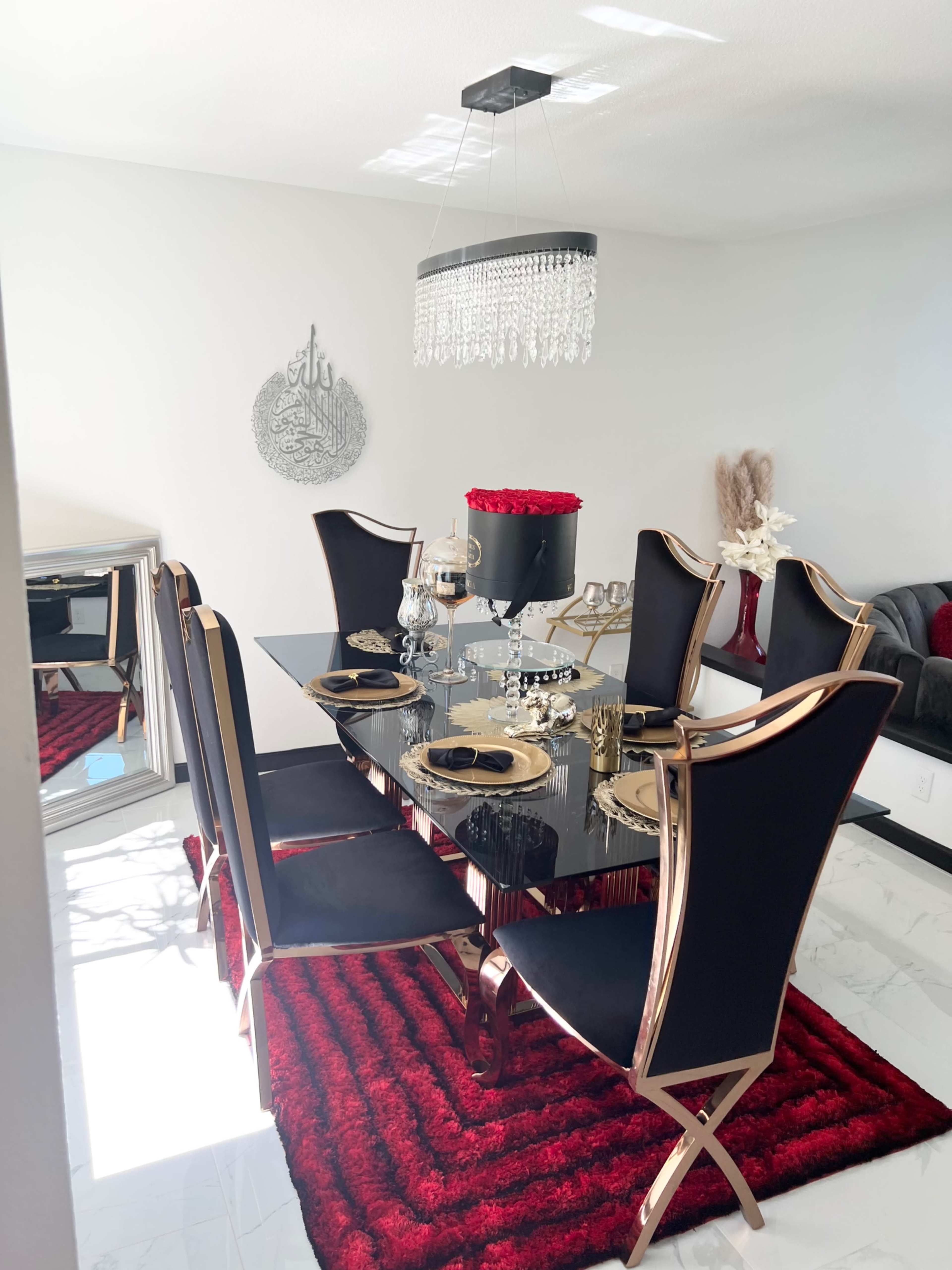 A modern dining room with a black glass table surrounded by six elegant chairs, a red area rug, and a chandelier hanging above.