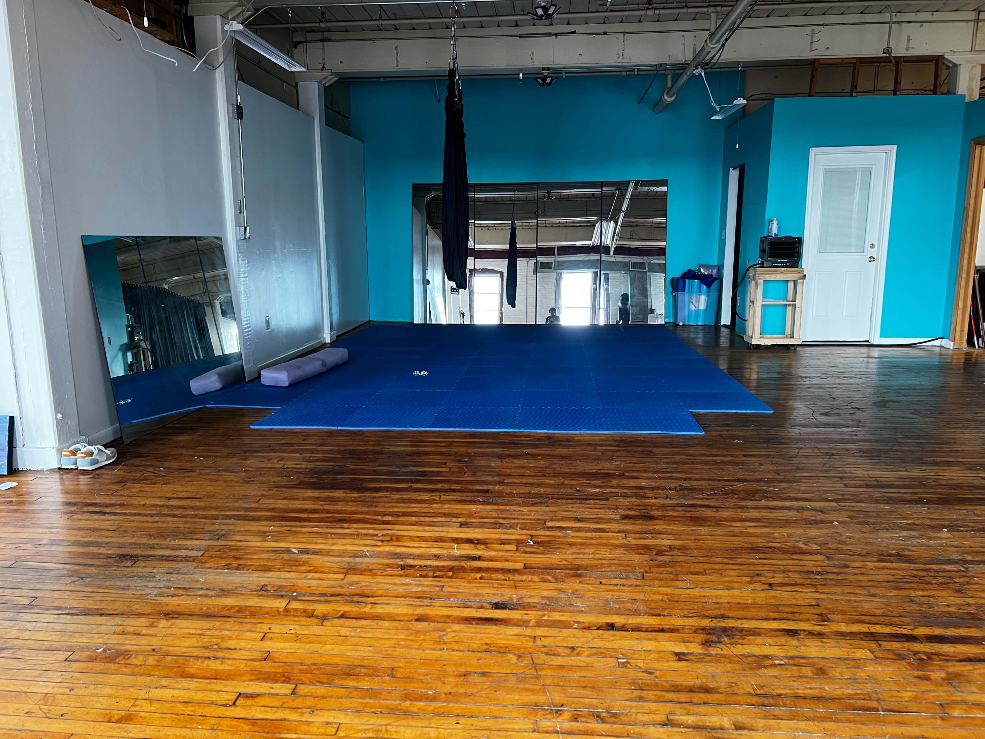 The image shows a spacious, brightly colored studio with blue walls and a wooden floor, featuring blue exercise mats and a large mirror on one side.