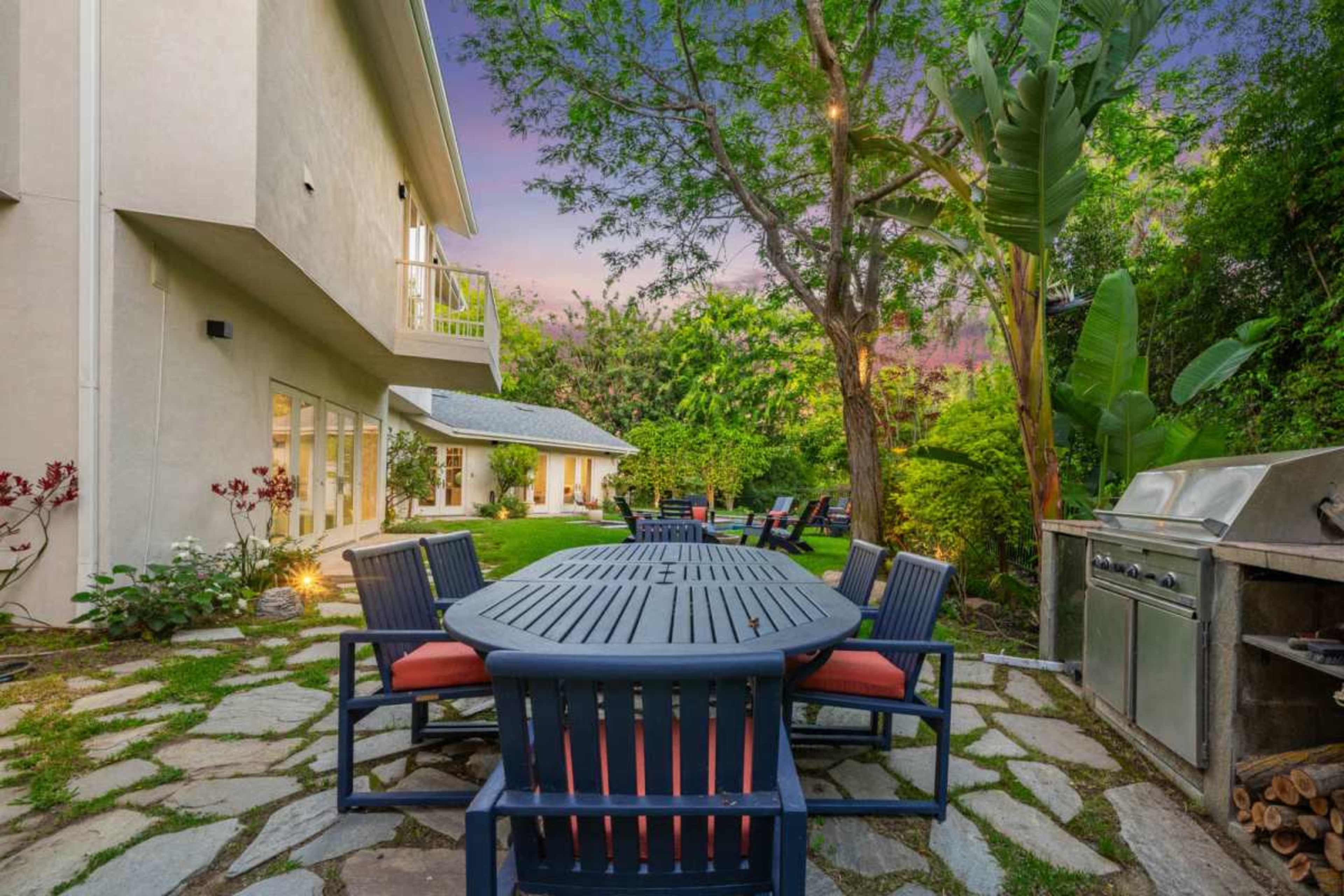 A stone patio features a large, dark wooden dining table surrounded by blue chairs, with a barbecue grill nearby and a lush garden in the background.