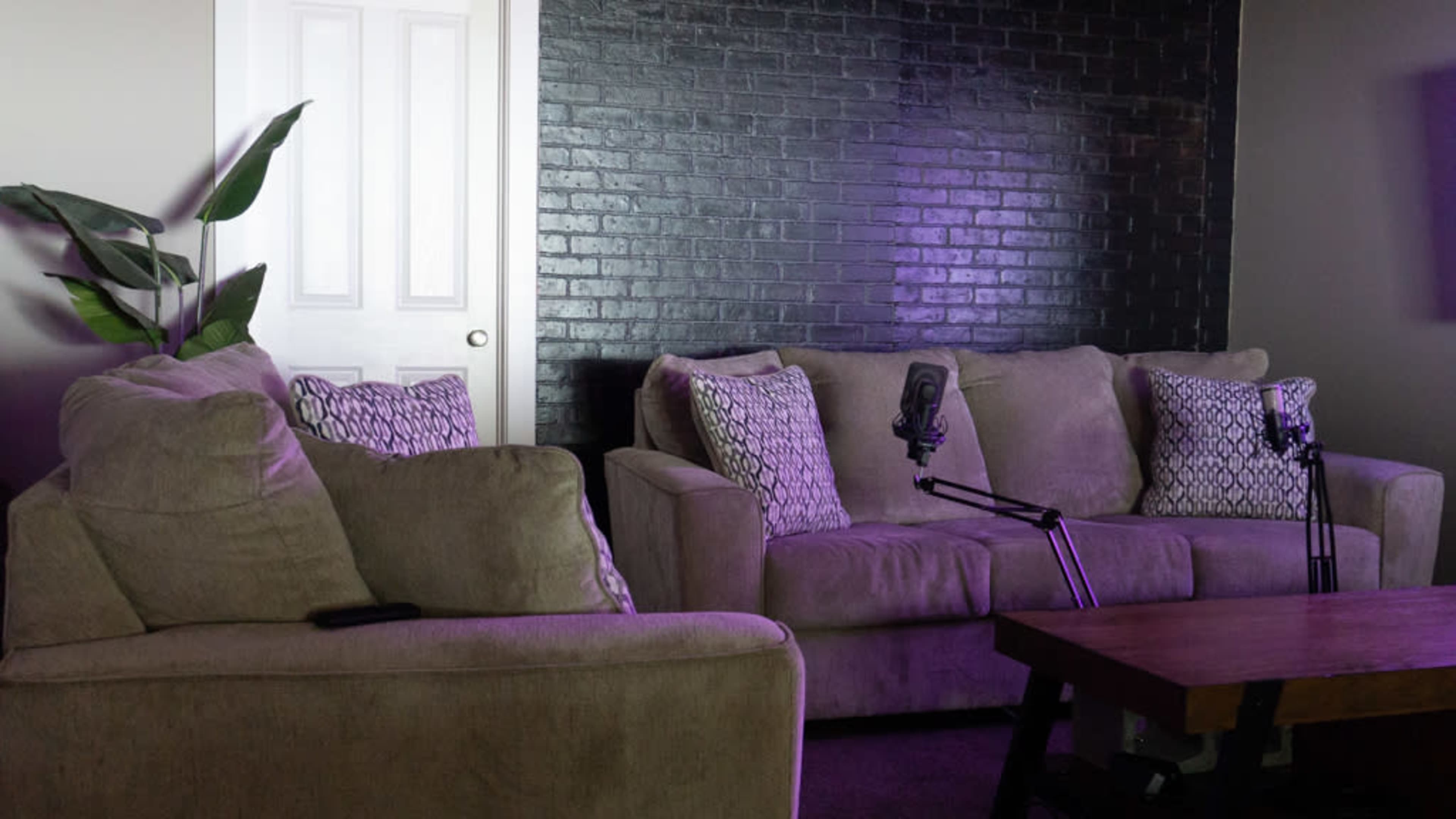 A living room features two beige sofas with decorative pillows, a wooden coffee table, and two microphones on stands, set against a dark brick wall and a closed white door.