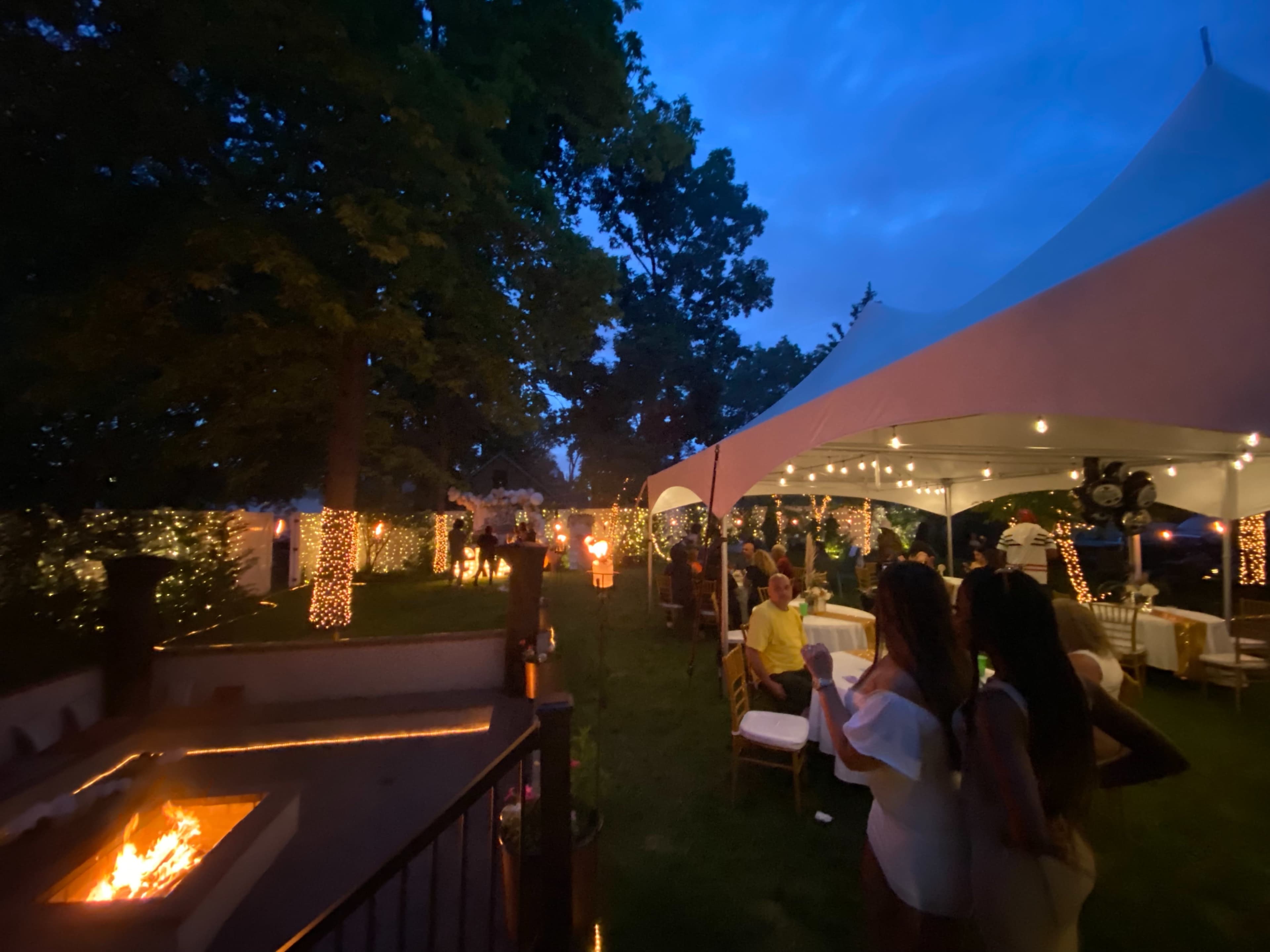 A festive outdoor gathering features a lit gazebo with string lights, a fire pit, and guests socializing under a large tent as dusk sets in.