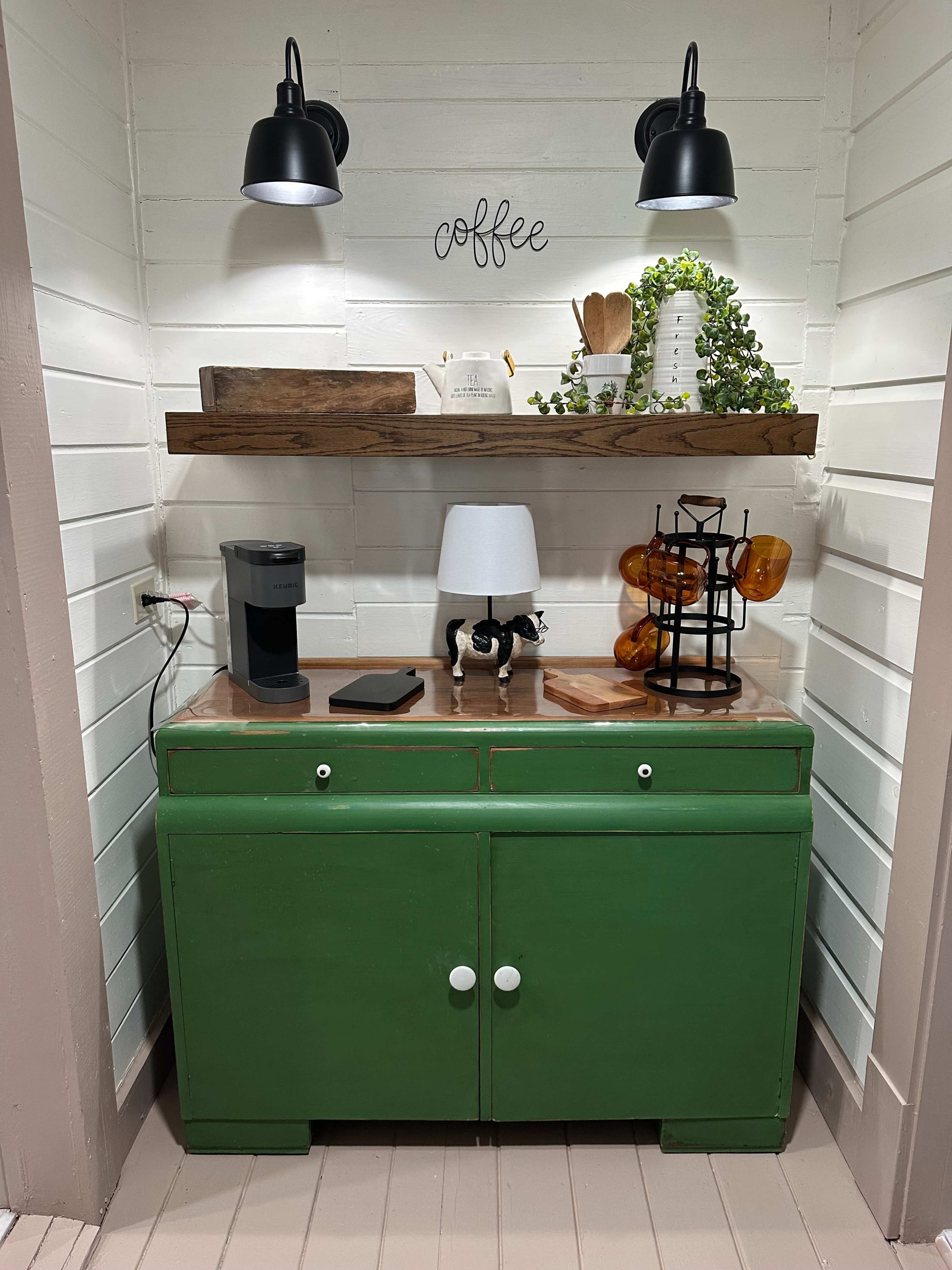 The image shows a green cabinet under a wooden shelf, adorned with a coffee maker, lamp, decorative items, and a wire stand holding amber-colored glass jars.