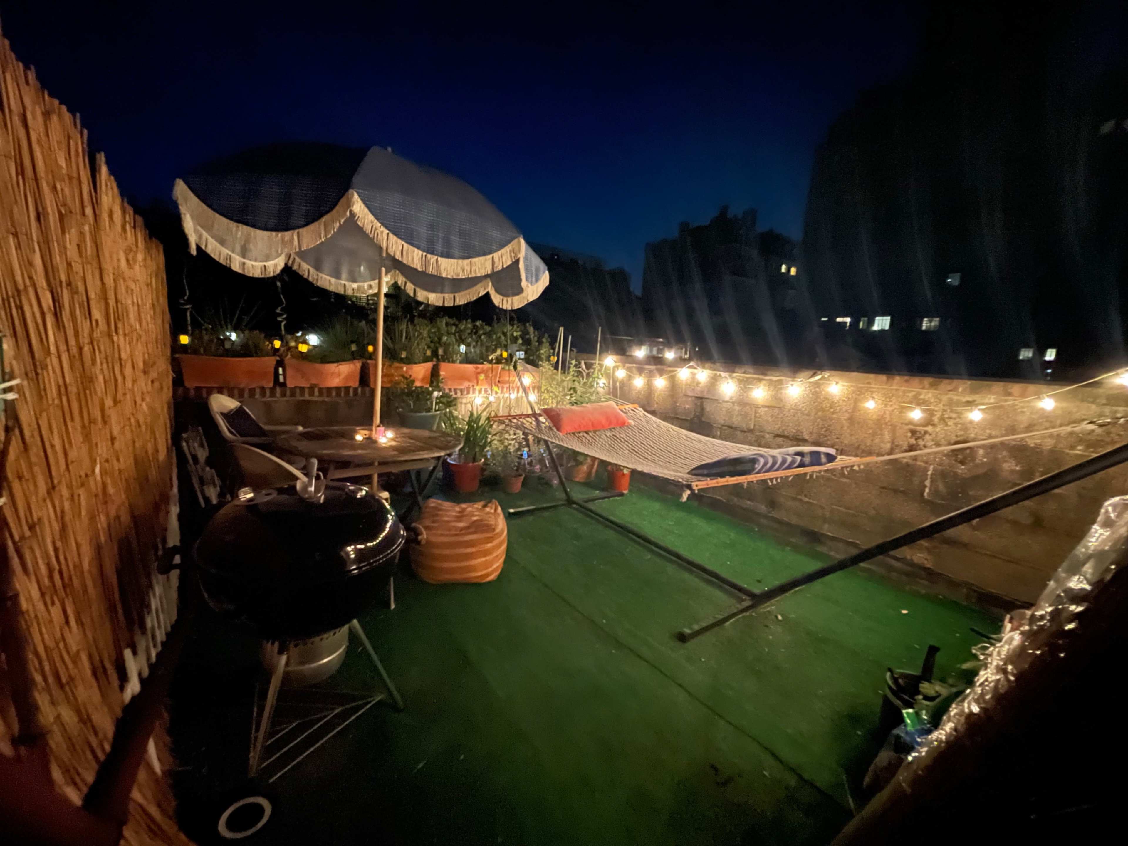 A rooftop terrace is illuminated by string lights and features a hammock, a grill, and potted plants under a large umbrella.