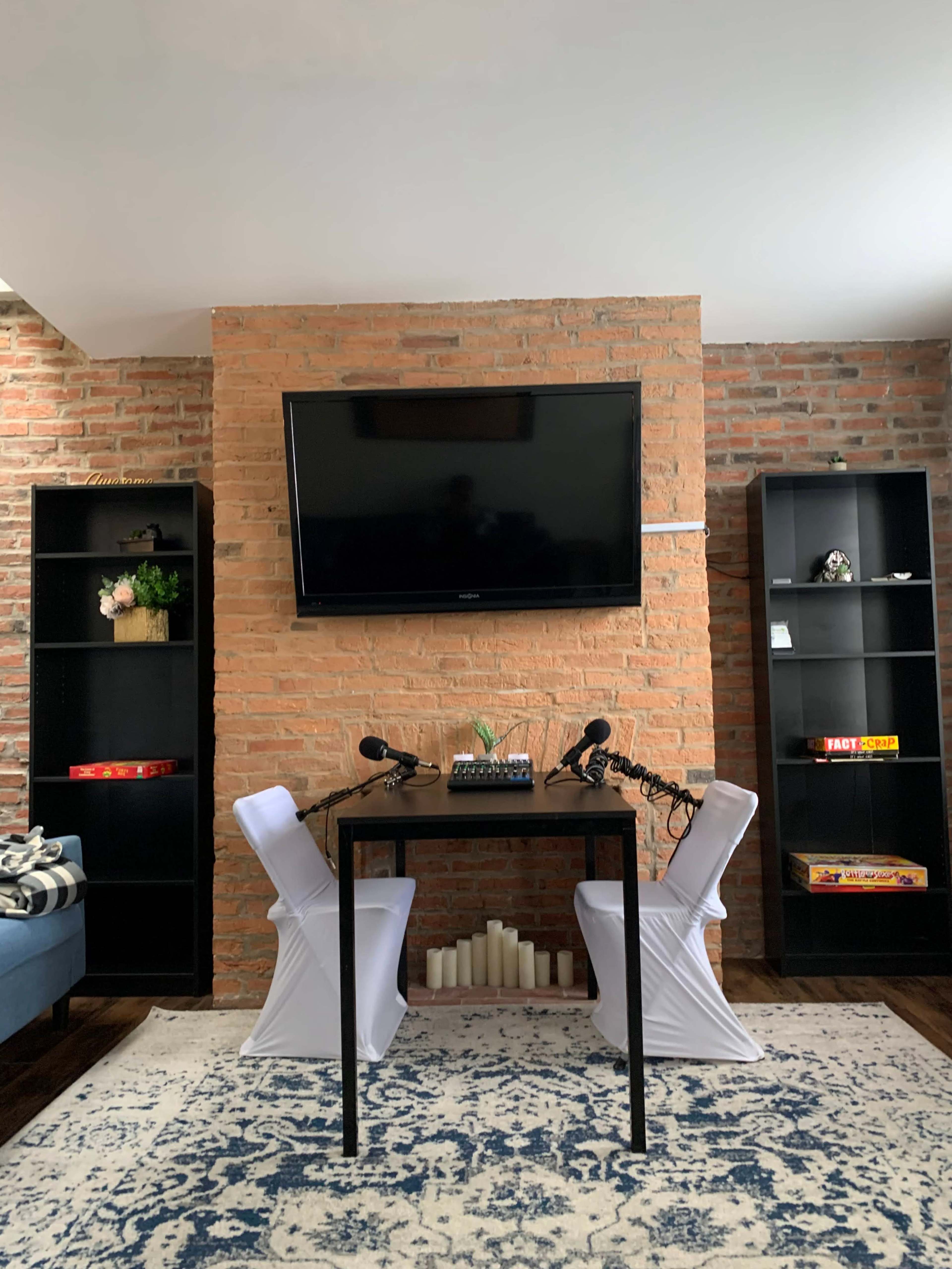A modern room features a black table with two microphones positioned on either side, against a backdrop of a brick wall and a large television mounted above, flanked by black shelving units.