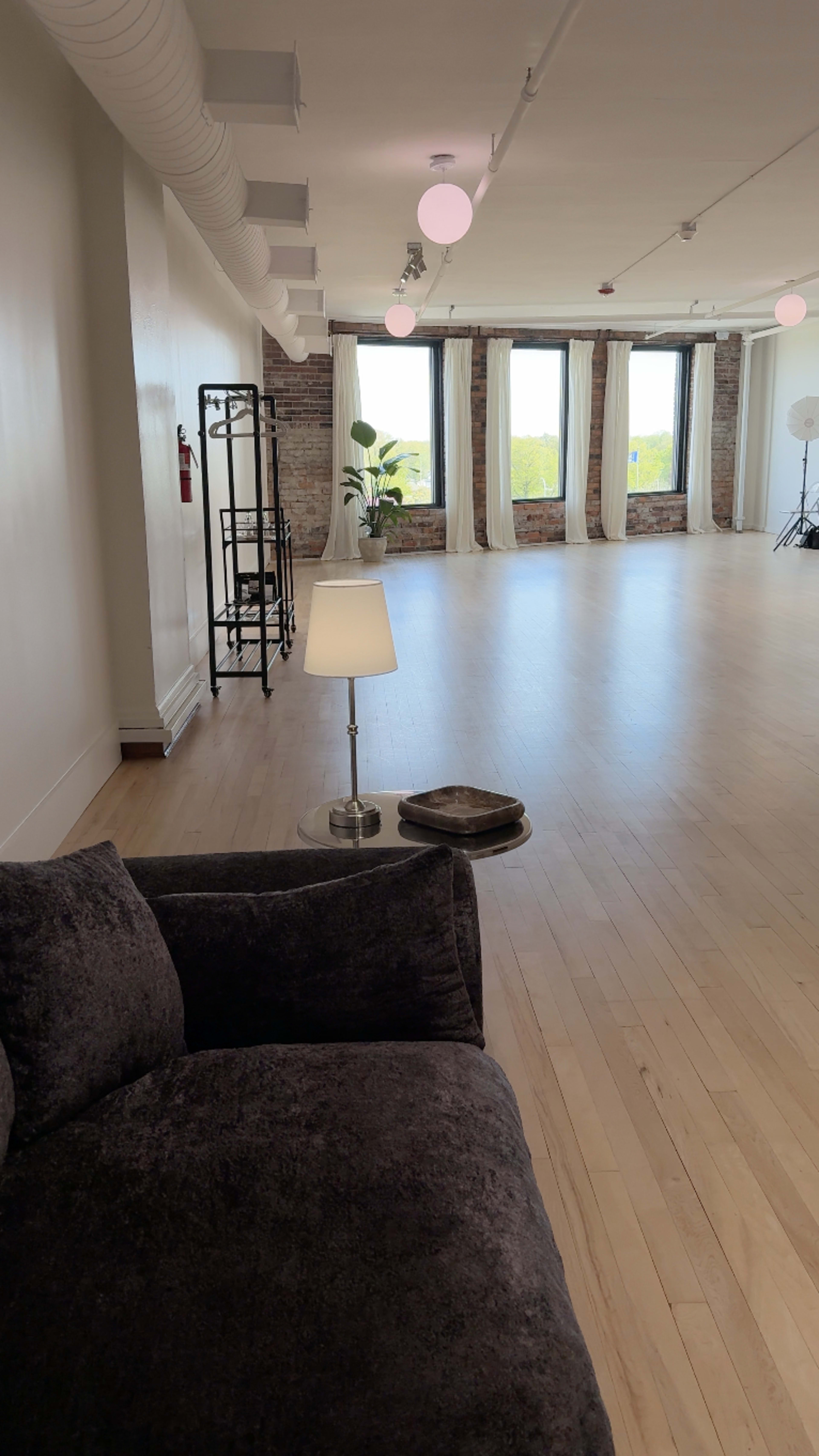 The image shows a spacious, well-lit room with wooden flooring, large windows, and a seating area featuring a gray couch and a lamp.