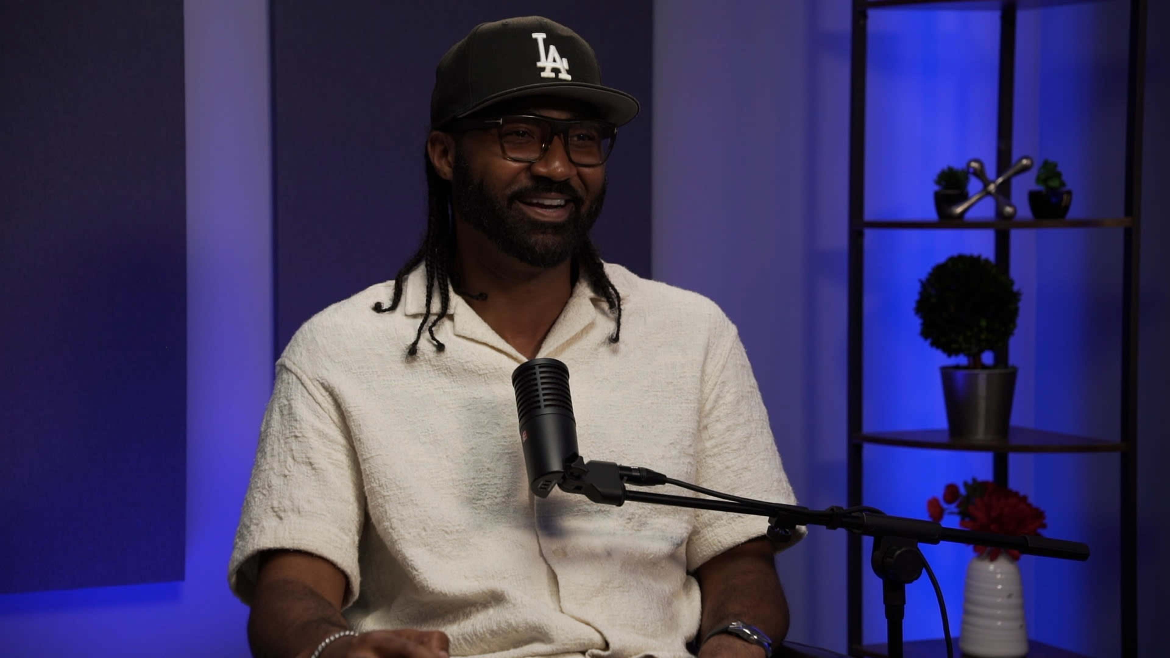 A man with braided hair, wearing a hat and glasses, is sitting in front of a microphone in a well-lit studio environment.