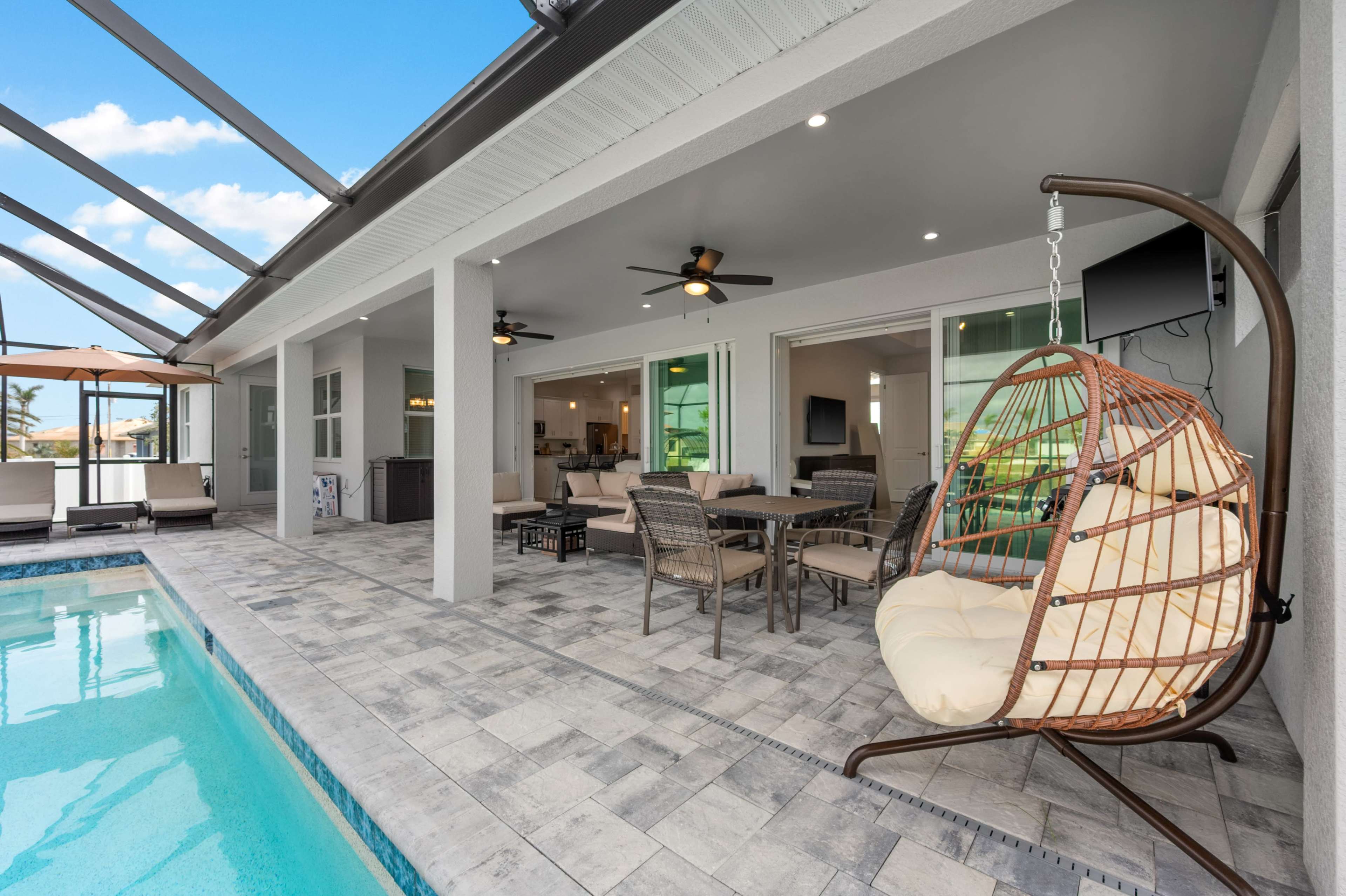 The image shows a modern outdoor patio area featuring a swinging chair, a dining table, and a swimming pool, all under a structured roof.