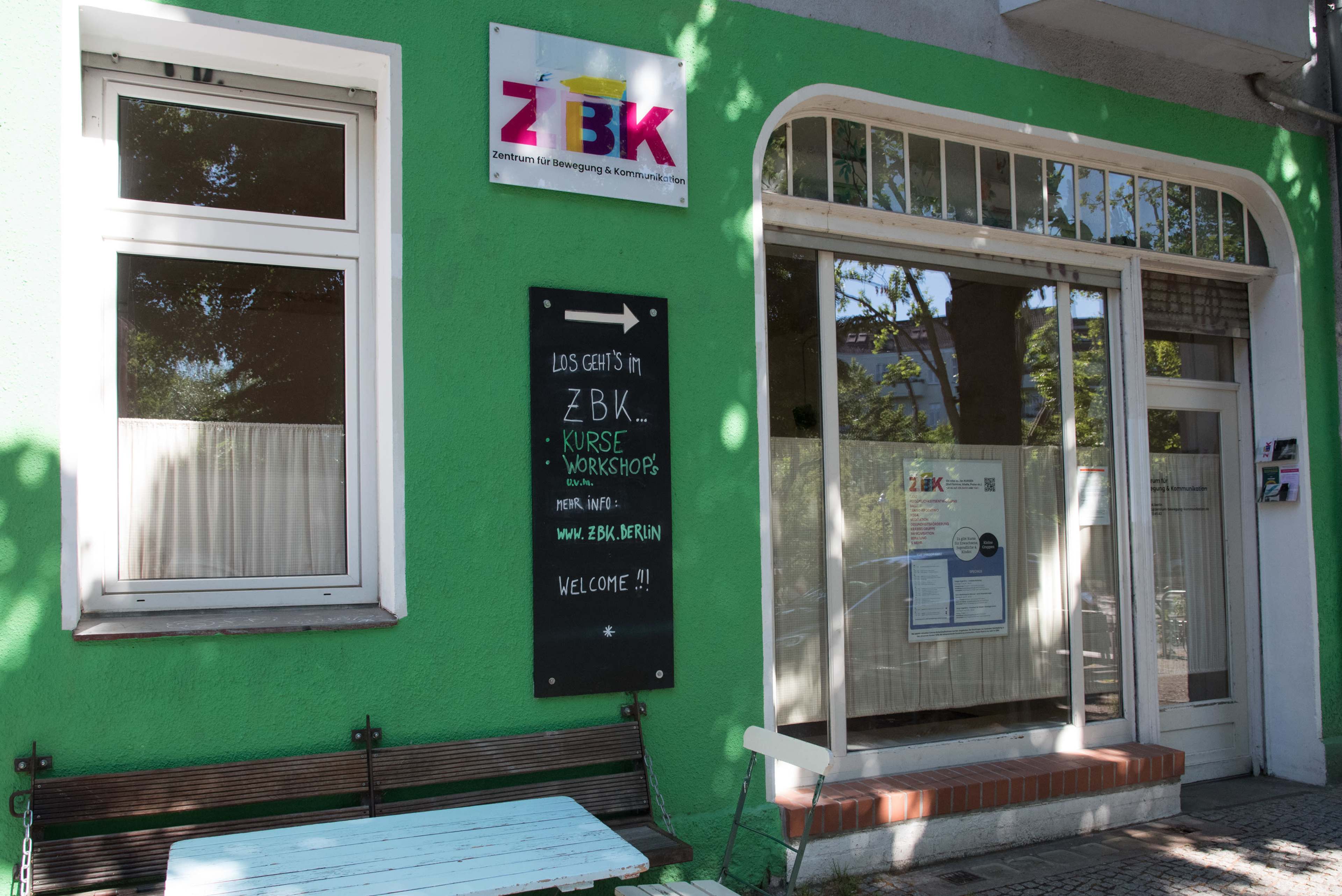 A storefront with a green exterior, featuring a sign that reads "ZBK" and a chalkboard displaying information about workshops and welcome messages.