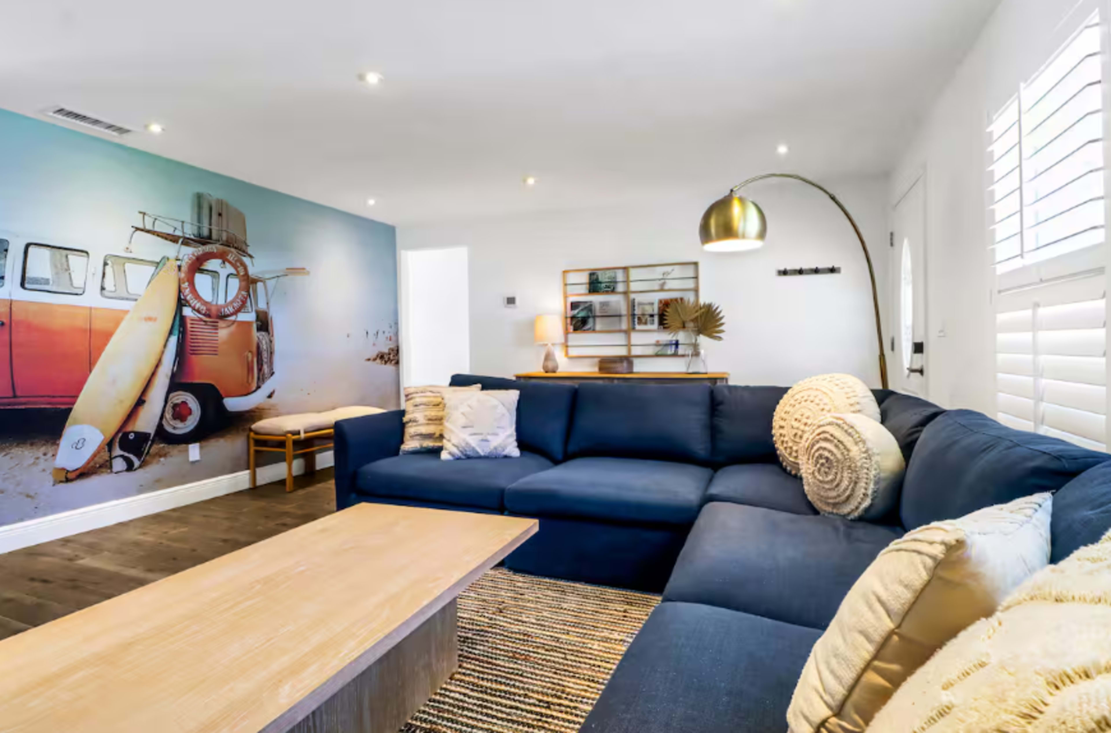 A cozy living room features a large blue sectional sofa, a light wooden coffee table, and a mural of a vintage van with a surfboard on a wall.