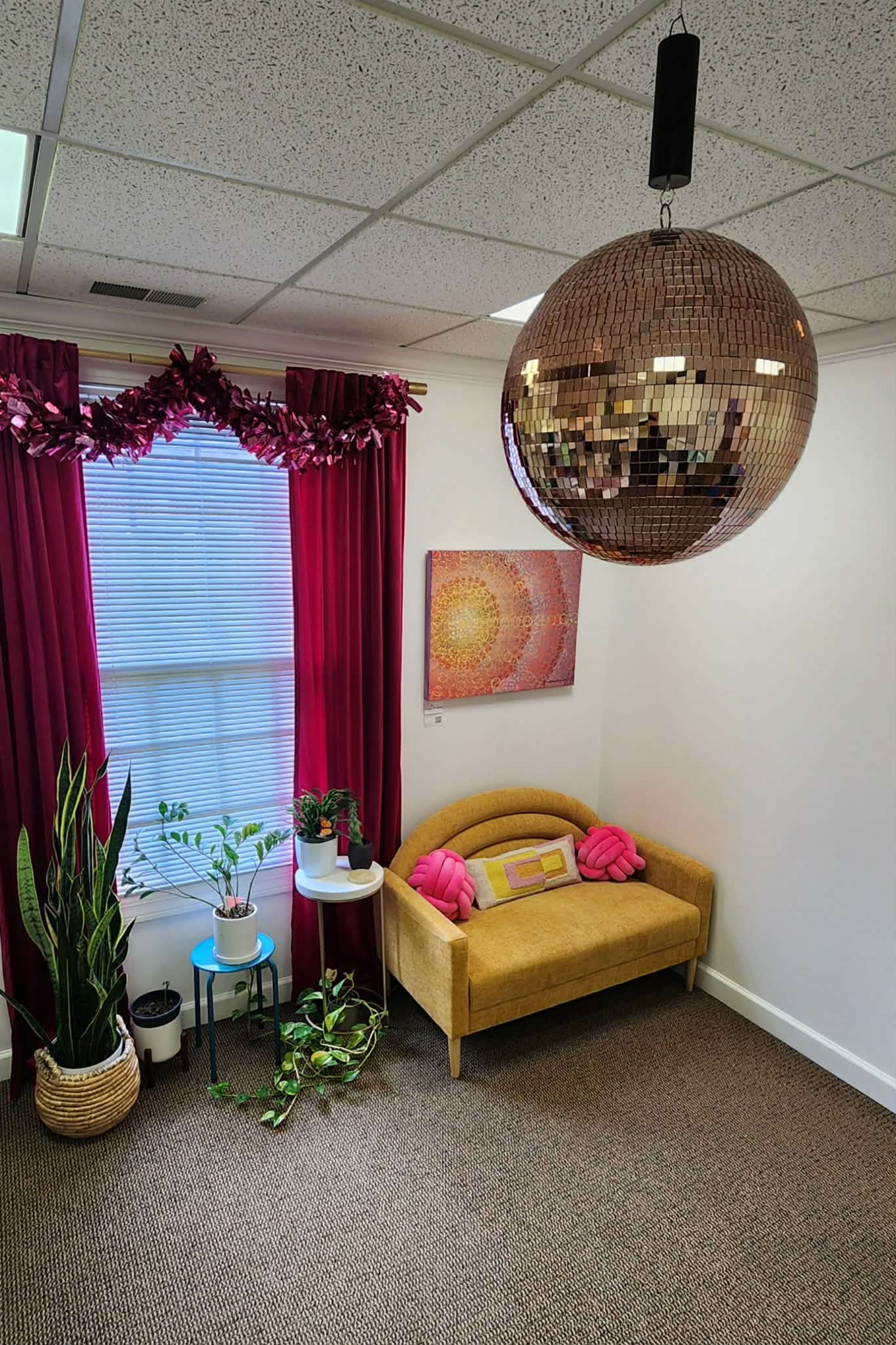 The image shows a cozy room with a yellow upholstered sofa, pink decorative pillows, a large disco ball hanging from the ceiling, and various plants placed in decorative pots.