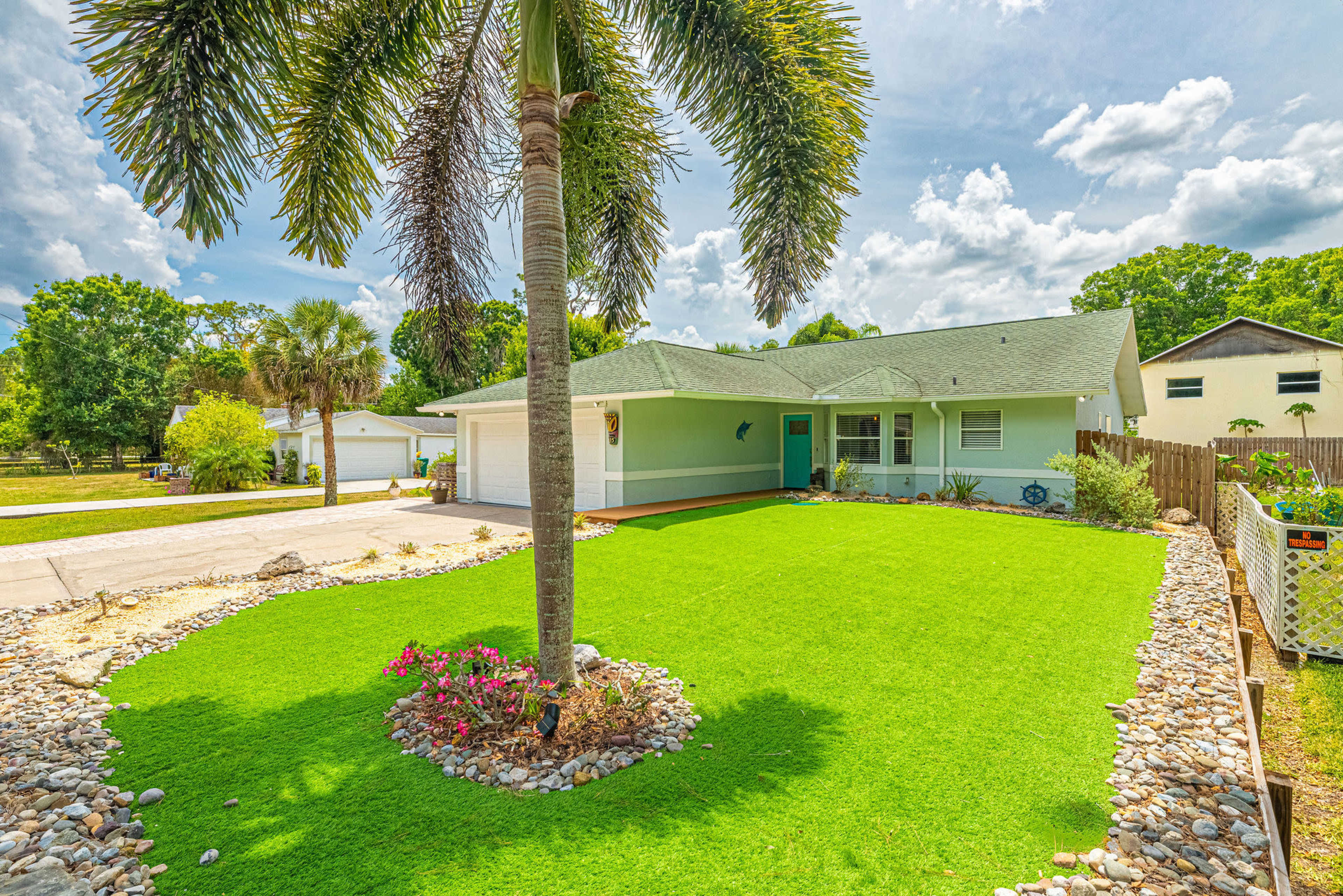 A single-story house with a light blue exterior is surrounded by well-maintained green lawn and decorative stones, featuring a palm tree in the foreground.