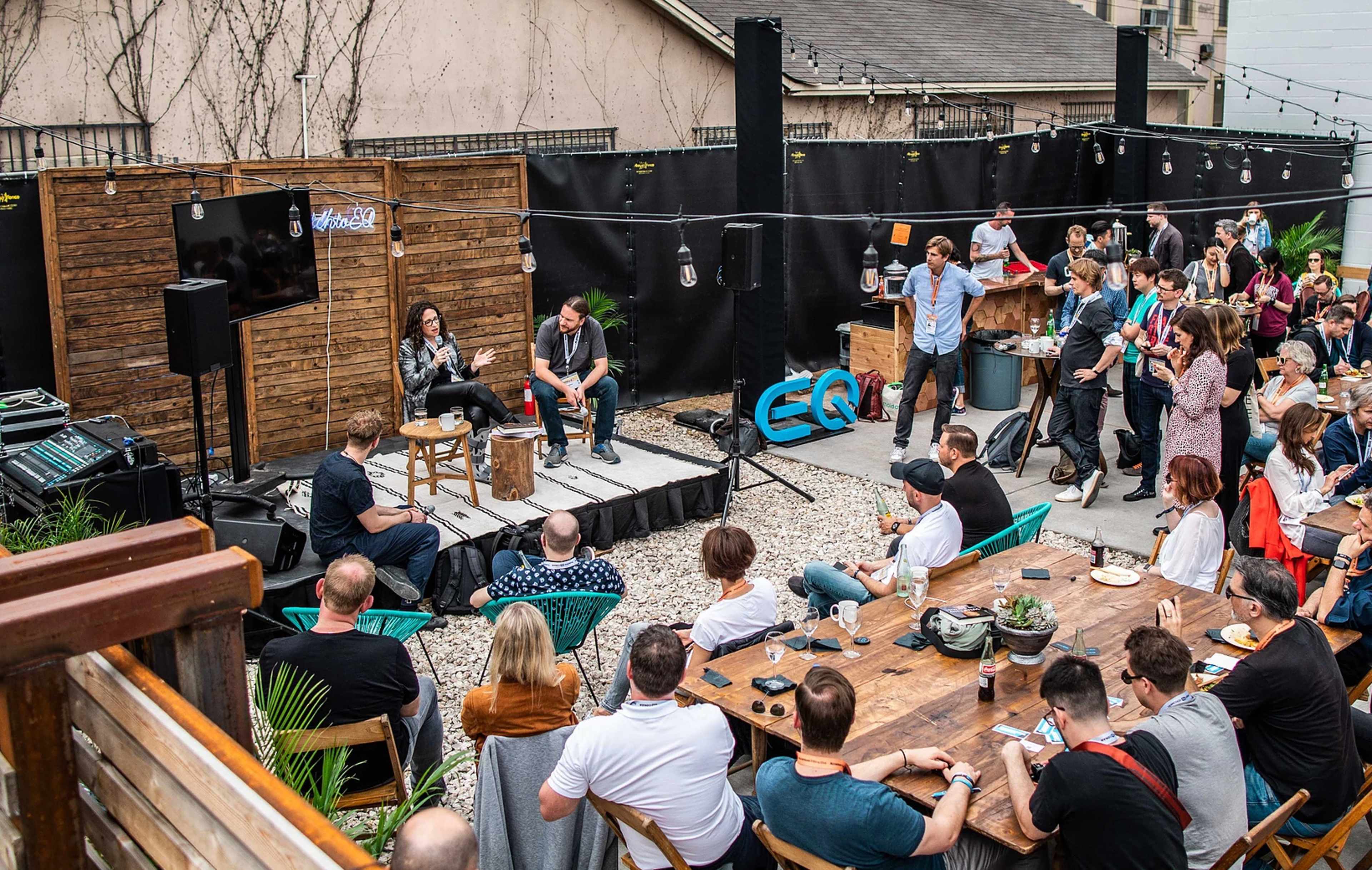 A group of people gathers outdoors for a panel discussion, with two speakers seated on a stage surrounded by an audience and various setup equipment.