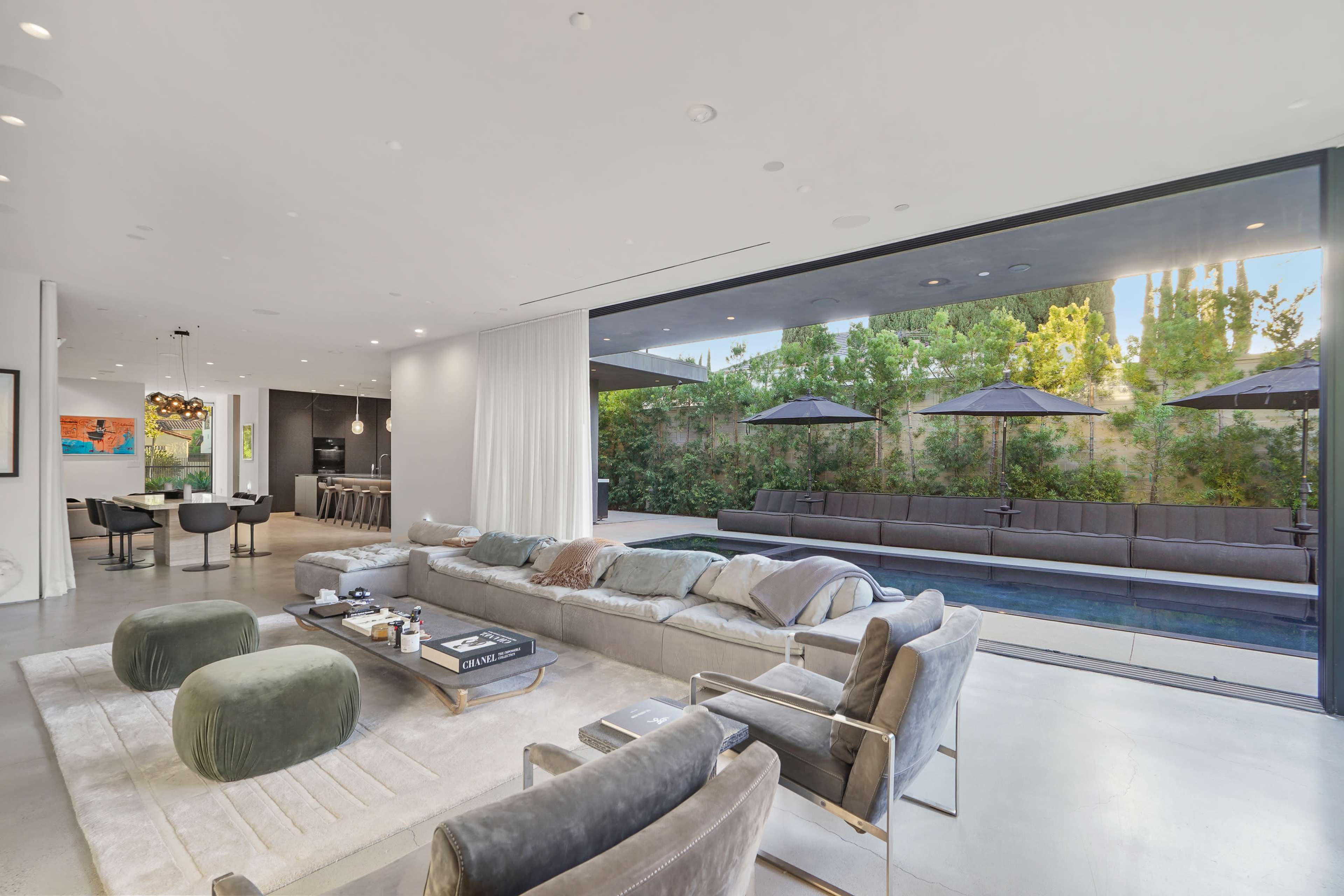 A modern living room features a spacious seating area that opens up to an outdoor patio with a pool and shaded lounge area.