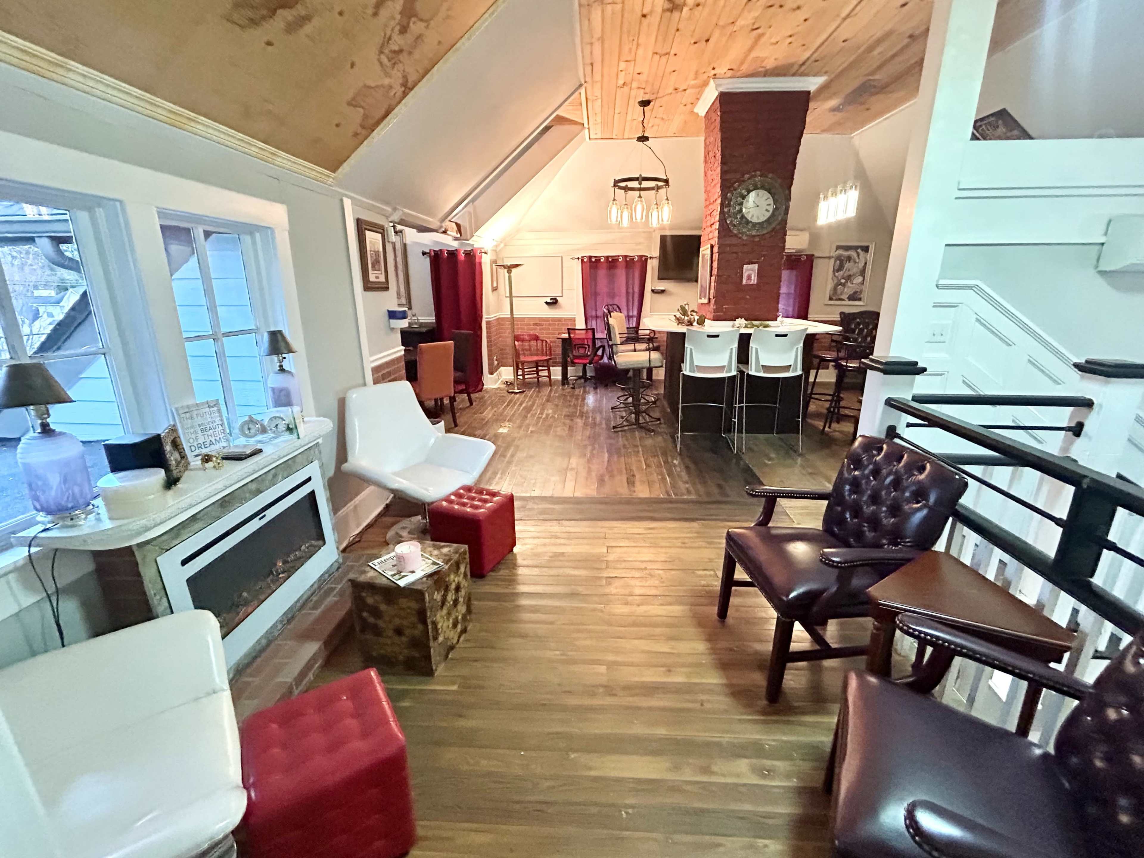 A cozy attic space with wooden floors, featuring a seating area with various chairs and a small fireplace, leading into a kitchen area with bar stools and a dining setup.
