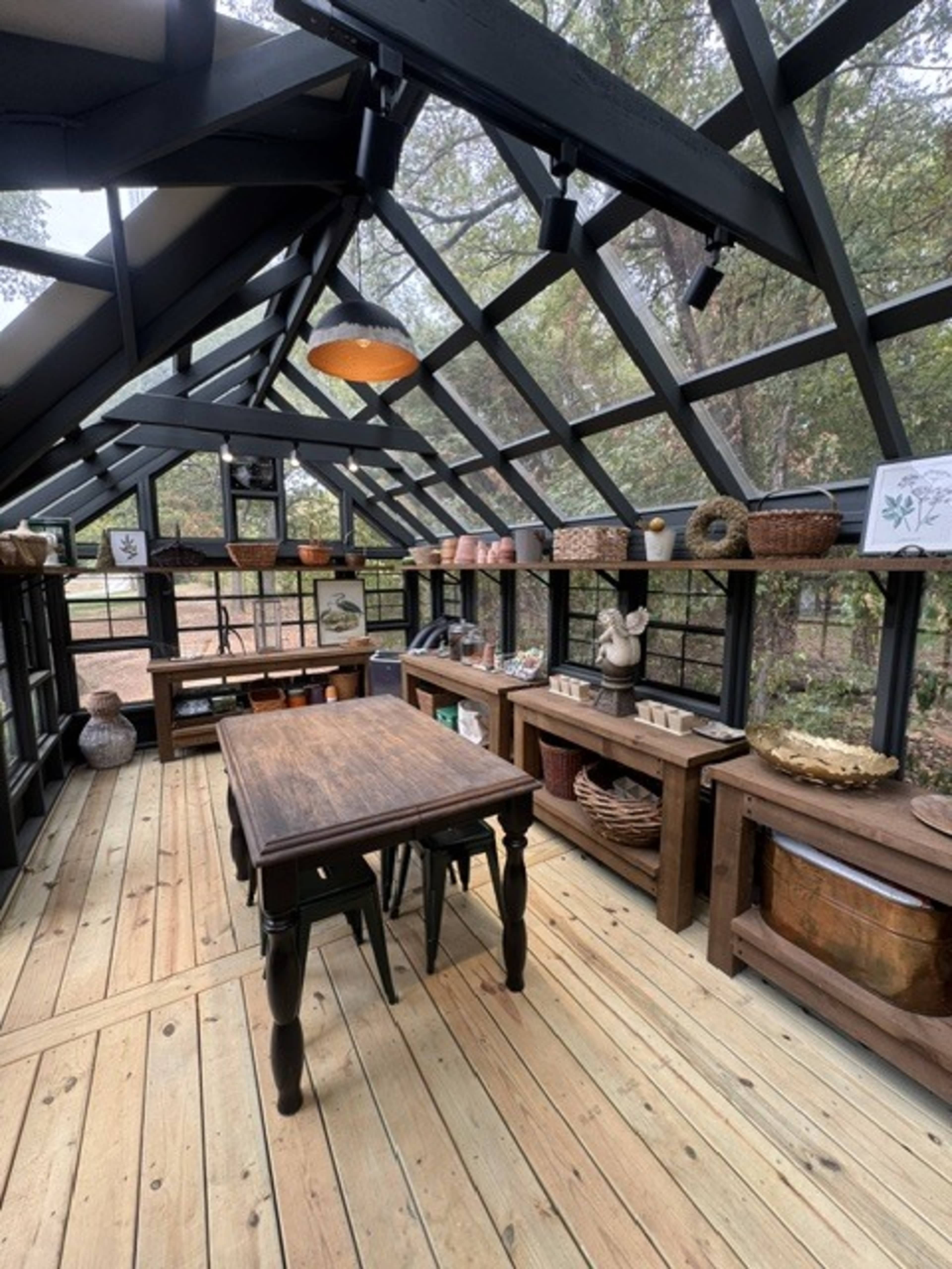 The image shows a spacious greenhouse with a glass roof, featuring a wooden table and shelves filled with various decorative baskets and pottery.