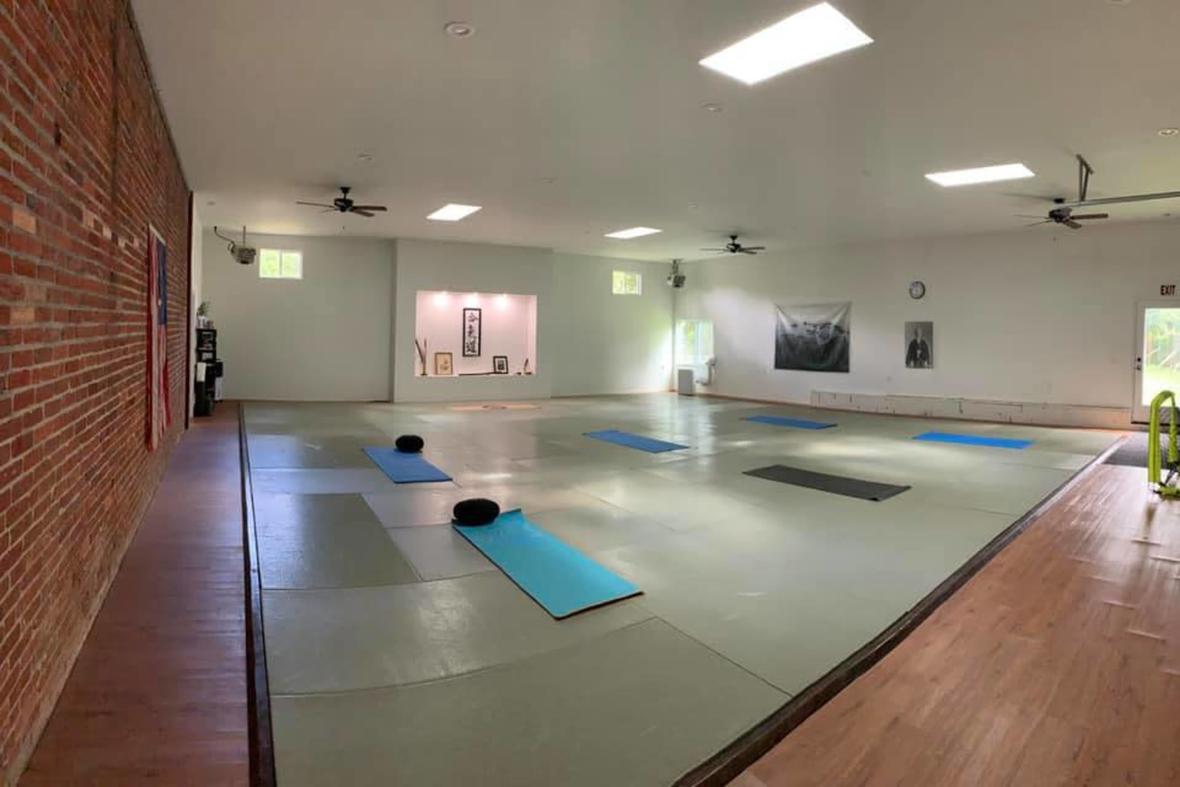The image shows a spacious, well-lit studio with wooden floors, a large mat area, and yoga mats arranged on the floor.