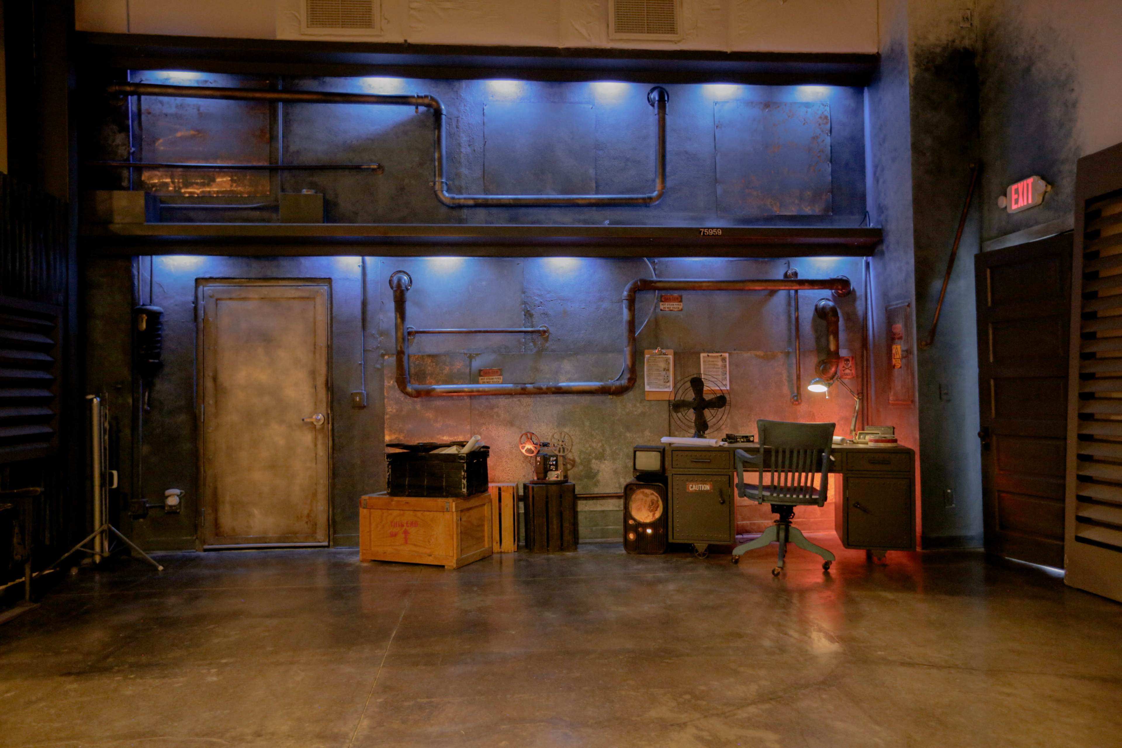 The image depicts an industrial-style room with a metallic wall, blue and red lighting, various pipes, a desk with a chair, and storage crates.