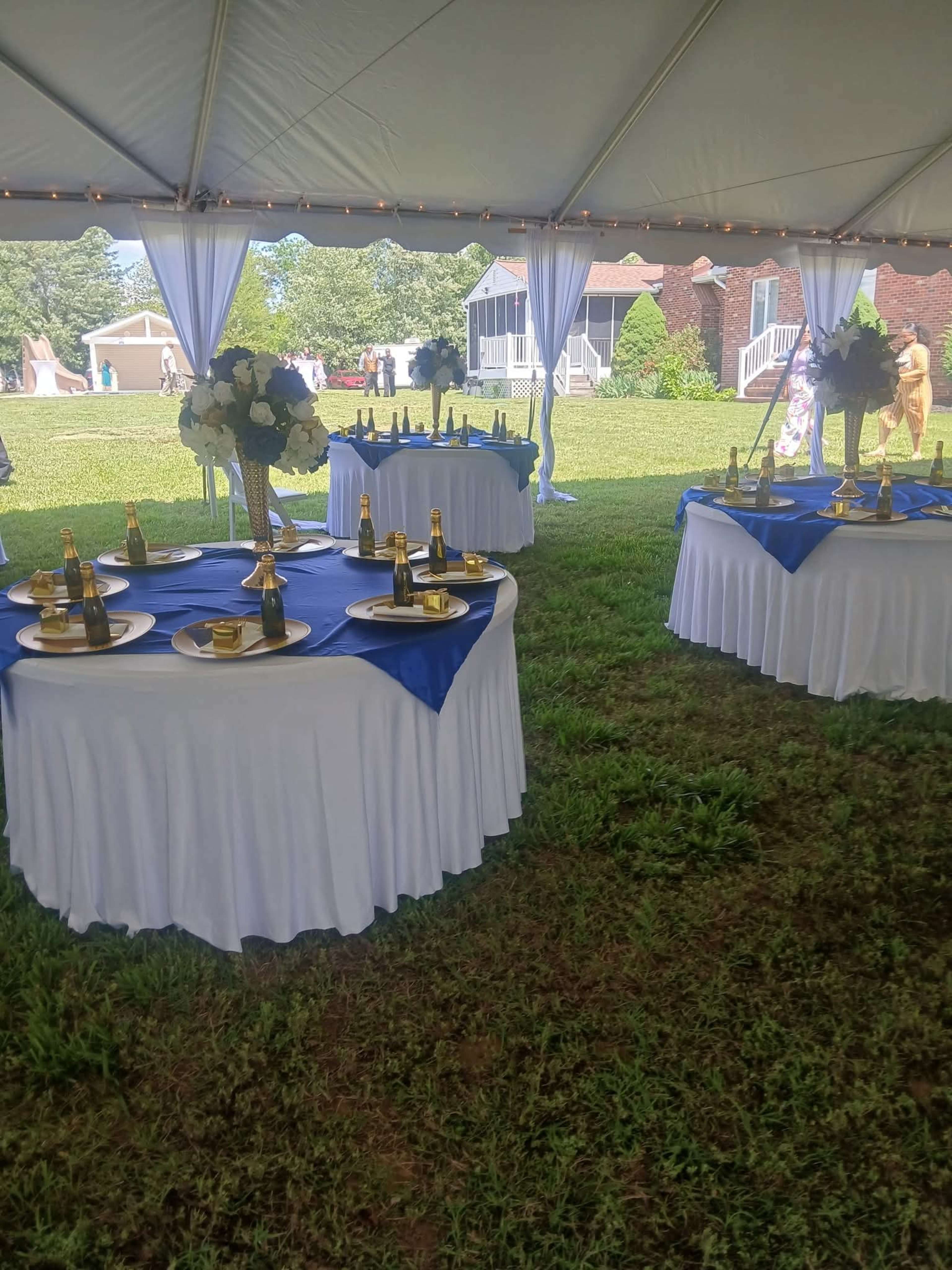 The image shows several round tables with white tablecloths and blue overlays set up under a tent, each adorned with champagne bottles and floral centerpieces on a grassy area.