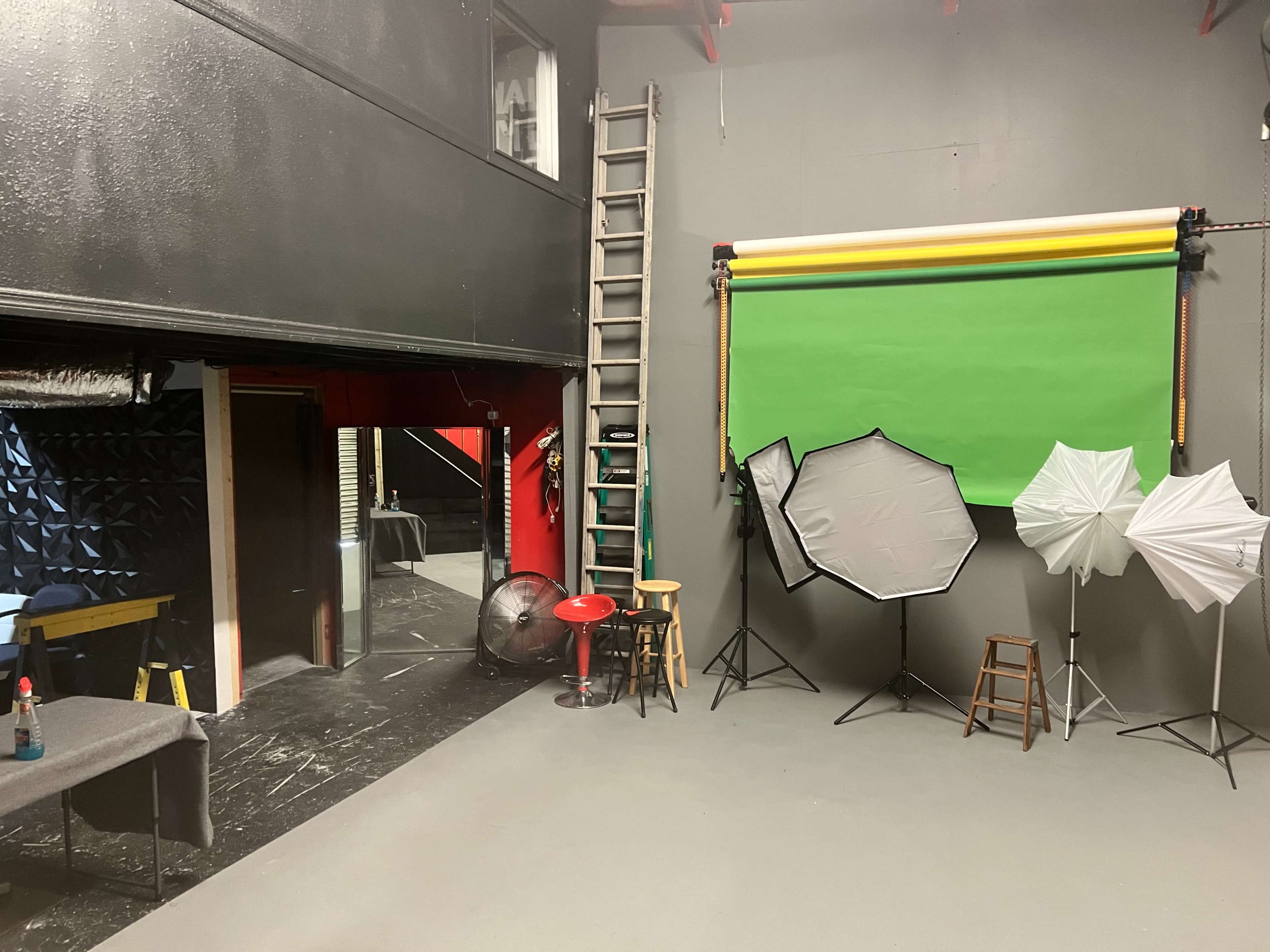 A photography studio with a green backdrop, several light diffusers, a ladder, and a fan stationed in the corner.