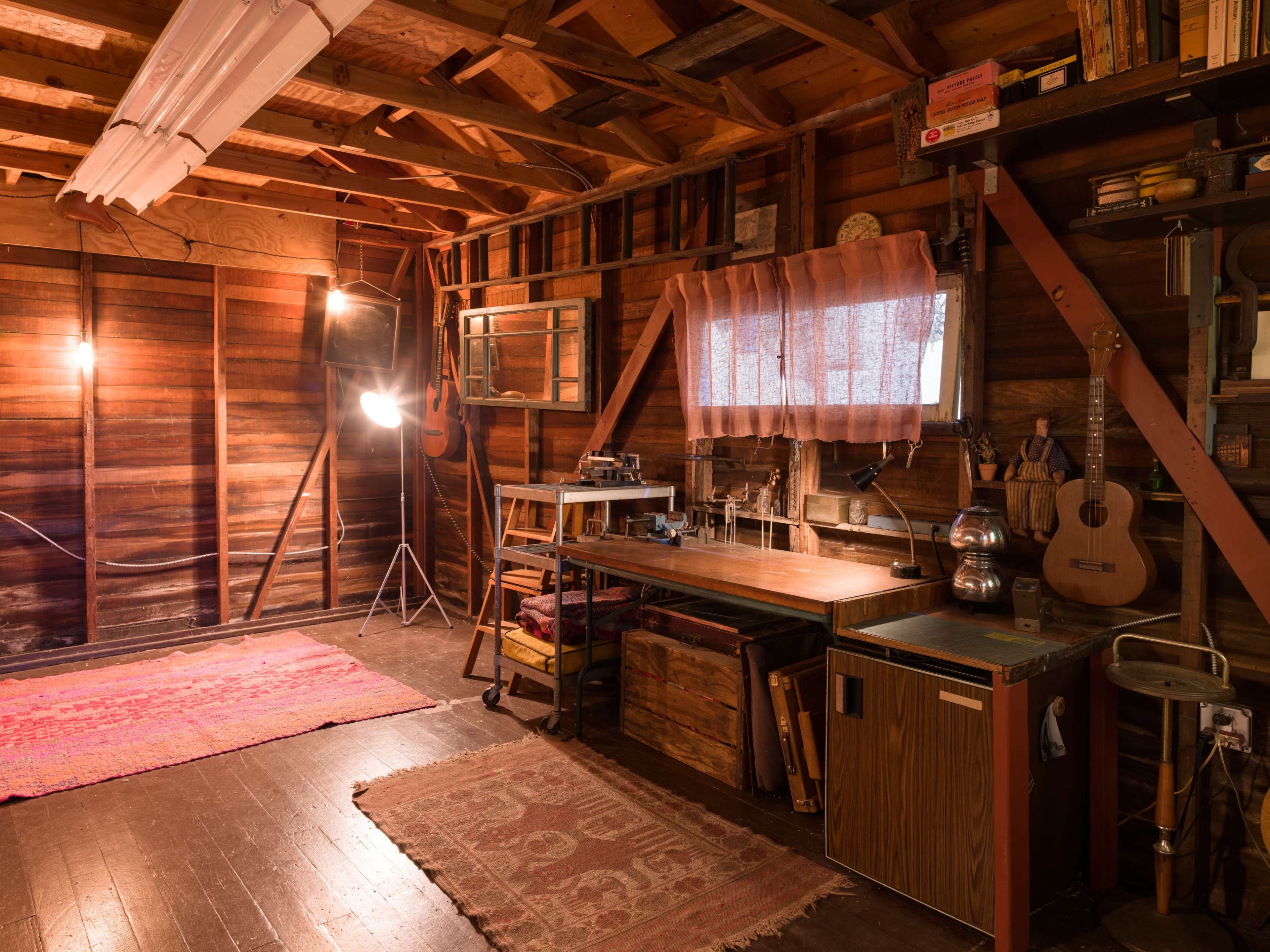 The image shows a rustic wooden interior of a workshop or studio with a worktable, shelves, and warm lighting.