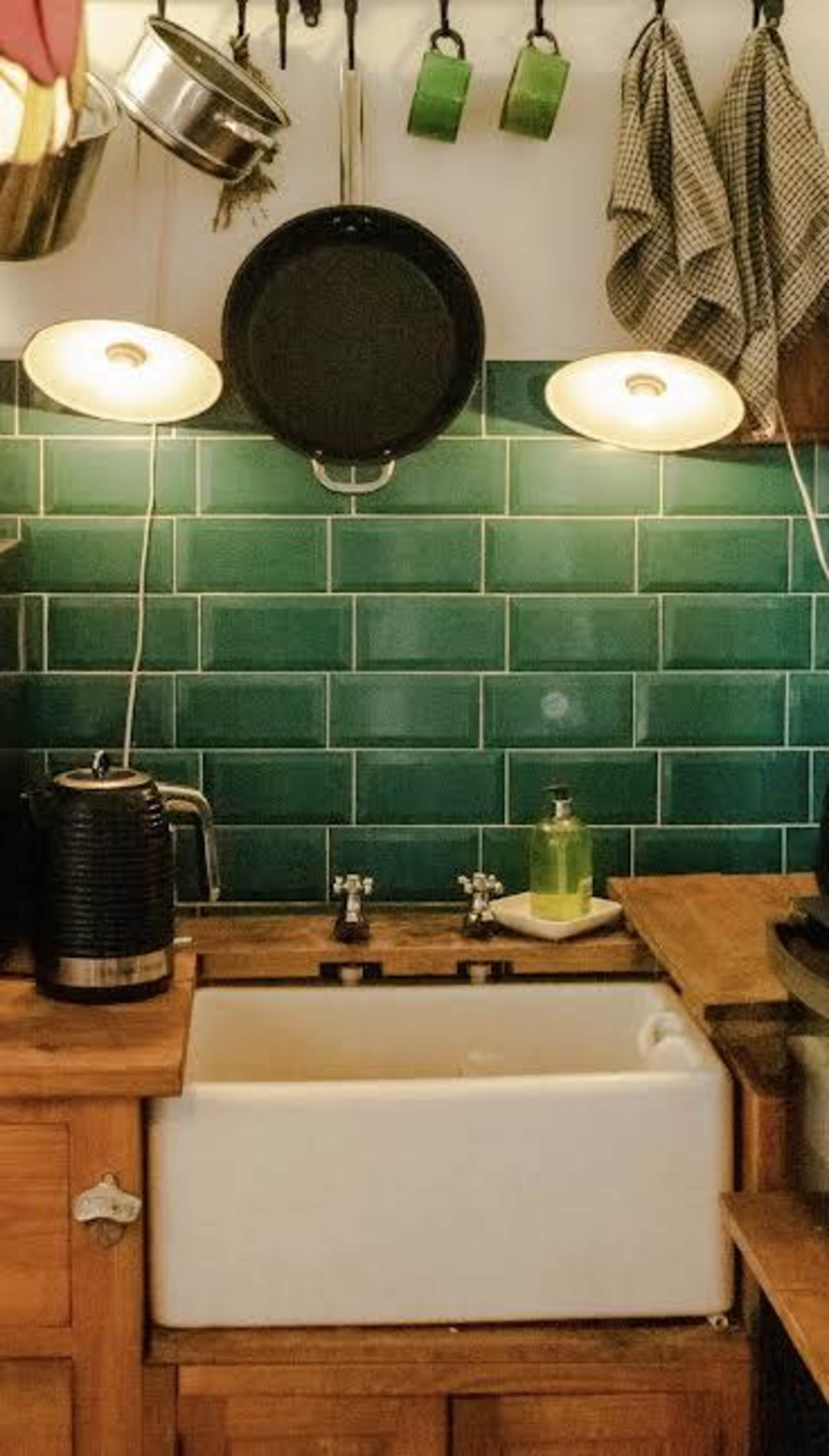 A kitchen with a white farmhouse sink, green tiled backsplash, and hanging cookware.