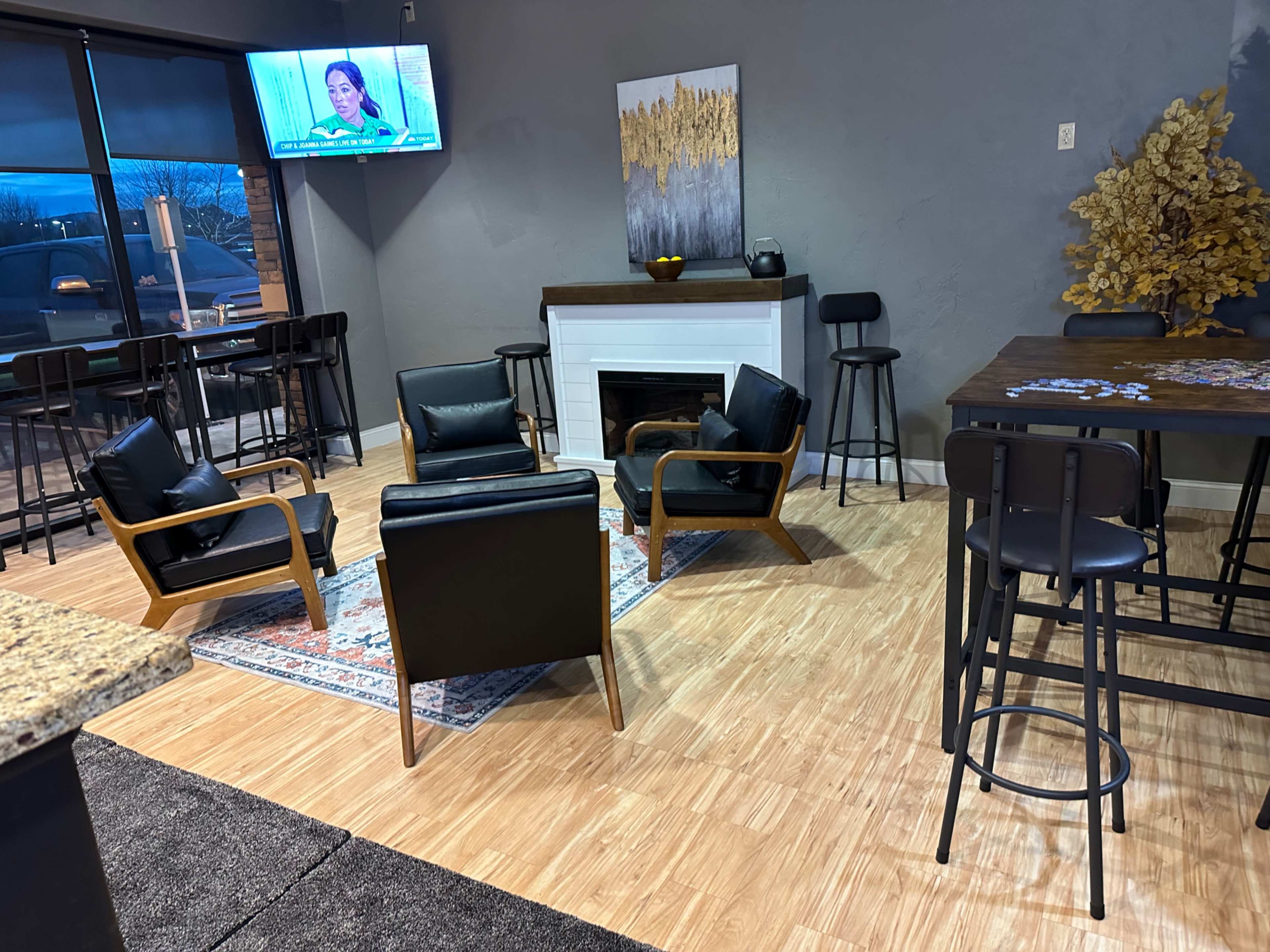 The scene features a cozy lounge area with four black chairs arranged around a small rug, a fireplace, and a bar table in the background, while a television displays a program on the wall.