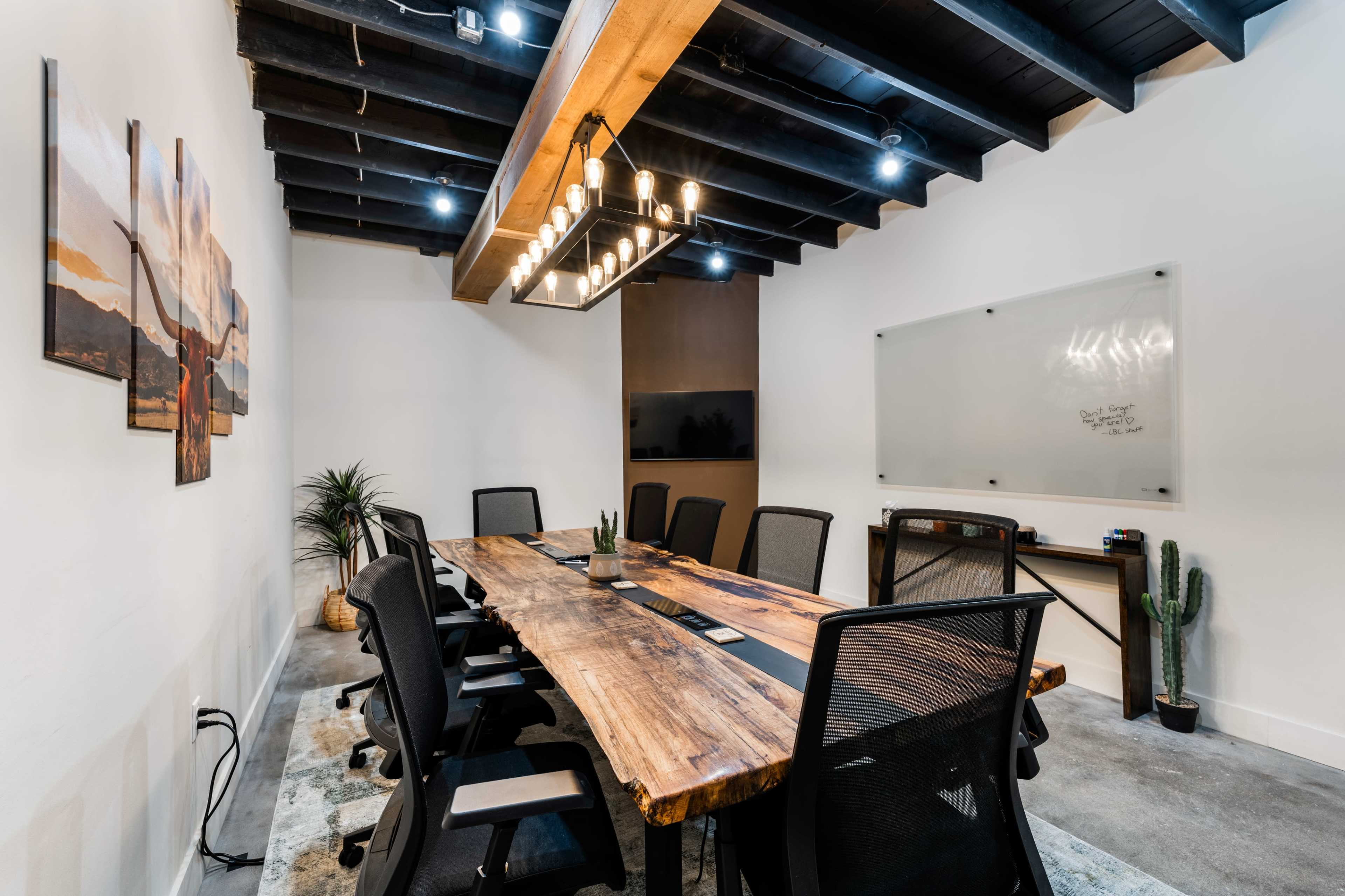 A modern conference room features a large wooden table surrounded by black chairs, with a whiteboard and television on the walls.