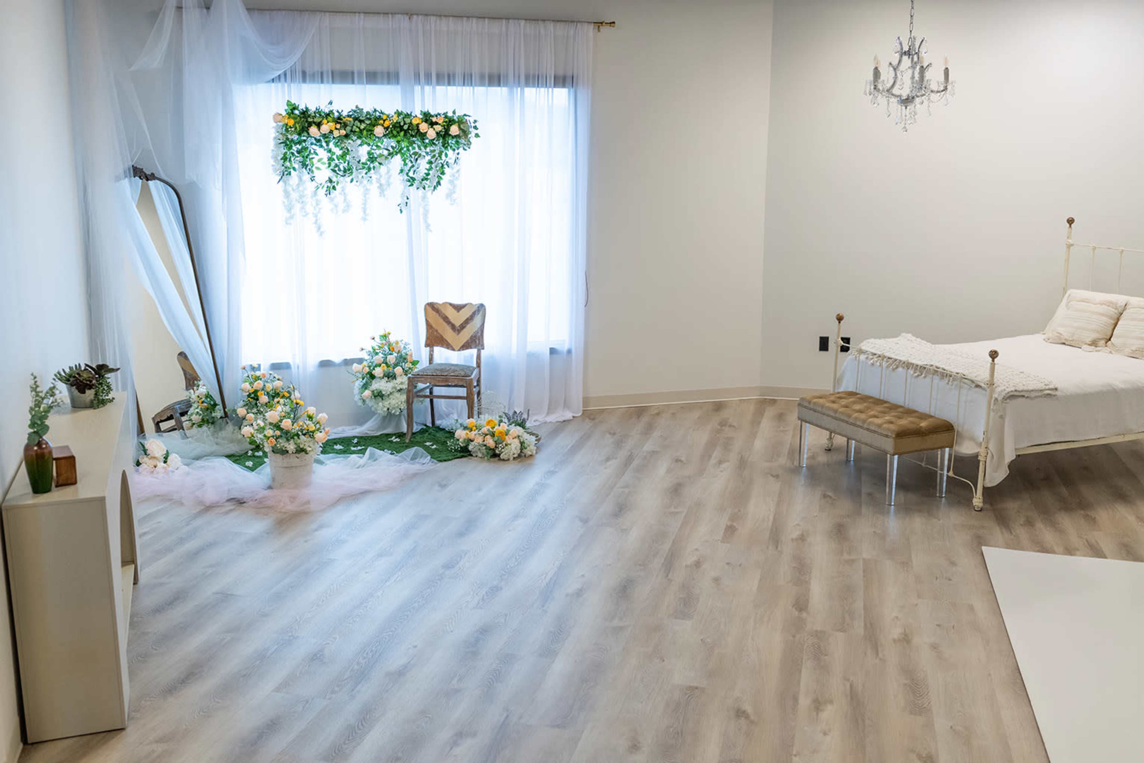 The image shows a bright room featuring a floral arrangement next to a window, a decorative chair, a bed, and a small bench on a wooden floor.