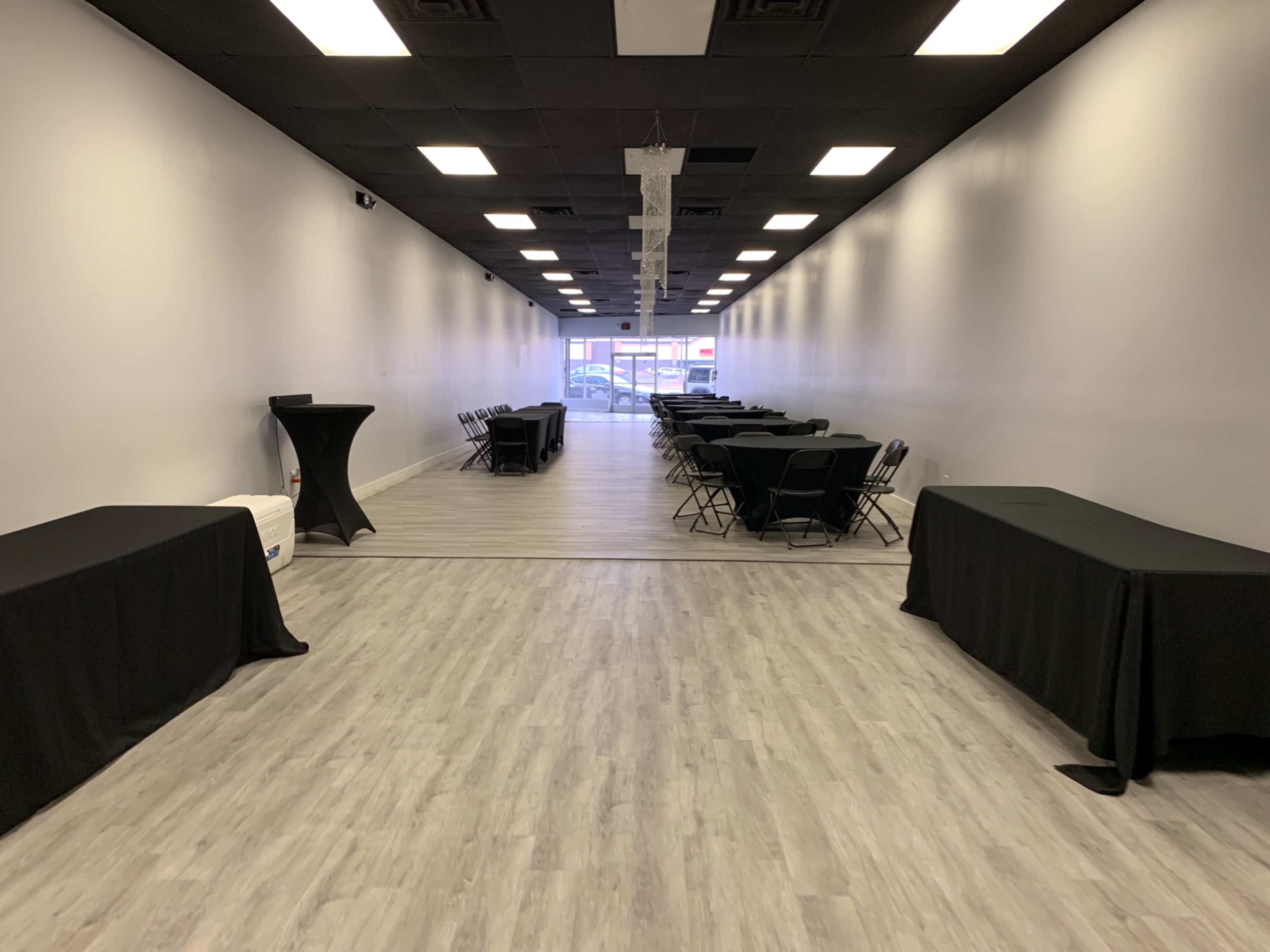 A spacious indoor venue with rows of black tables and chairs set up along a light-colored wooden floor, leading to a large entrance at the far end.