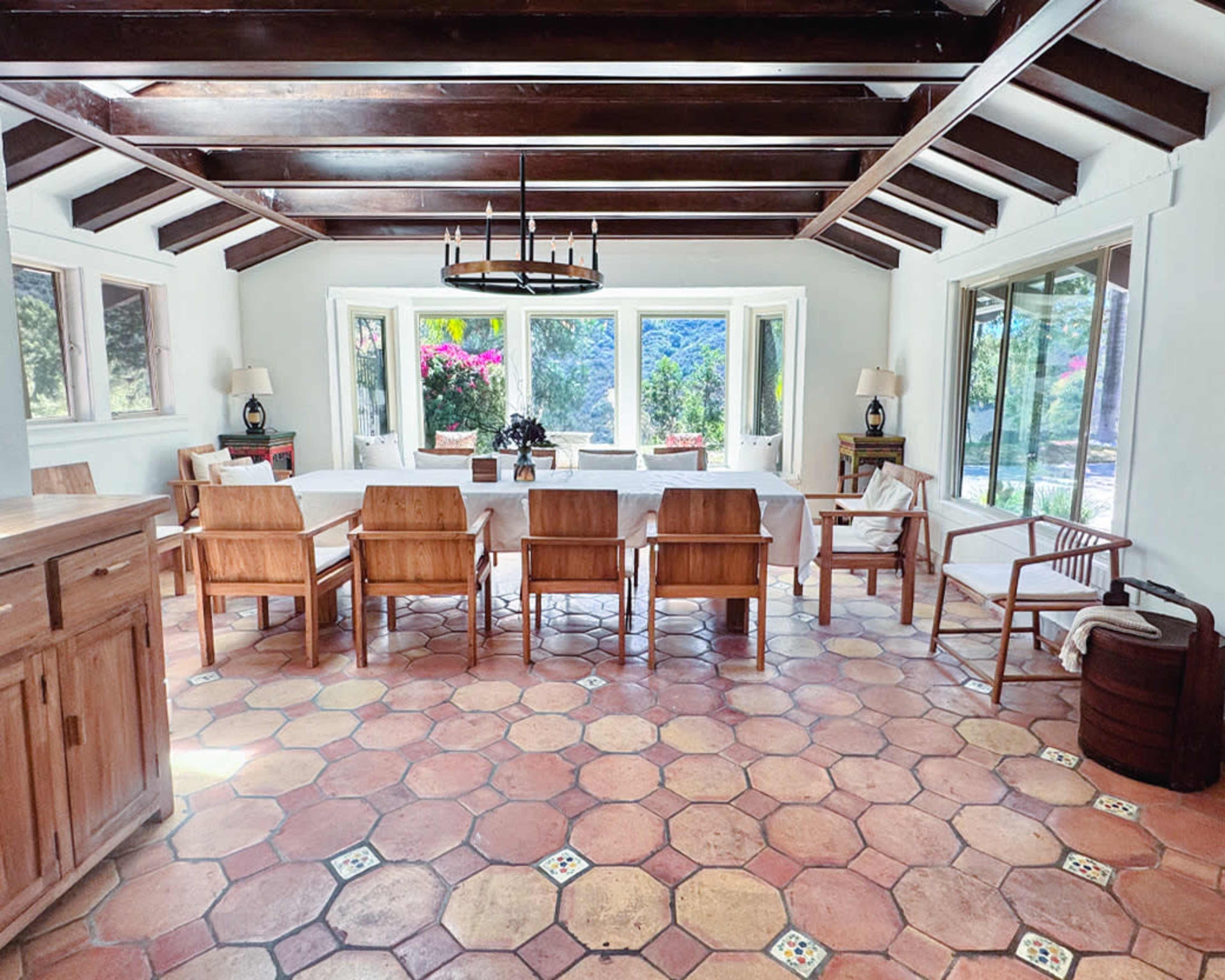 A dining room features a long table surrounded by wooden chairs, with large windows providing a view of greenery outside.