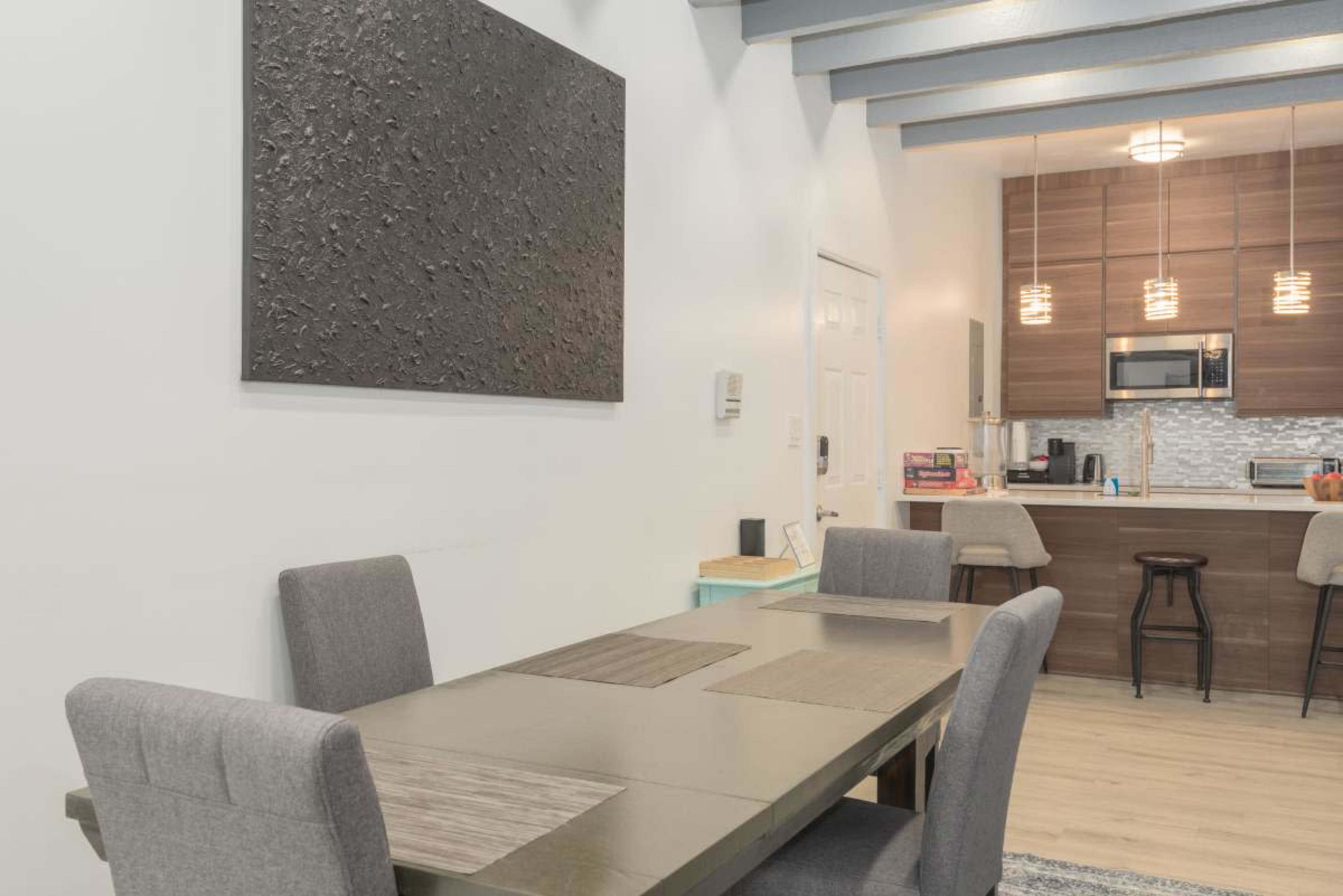 The image shows a modern dining area with a rectangular table and four gray chairs, featuring a textured wall art piece and a kitchen in the background.