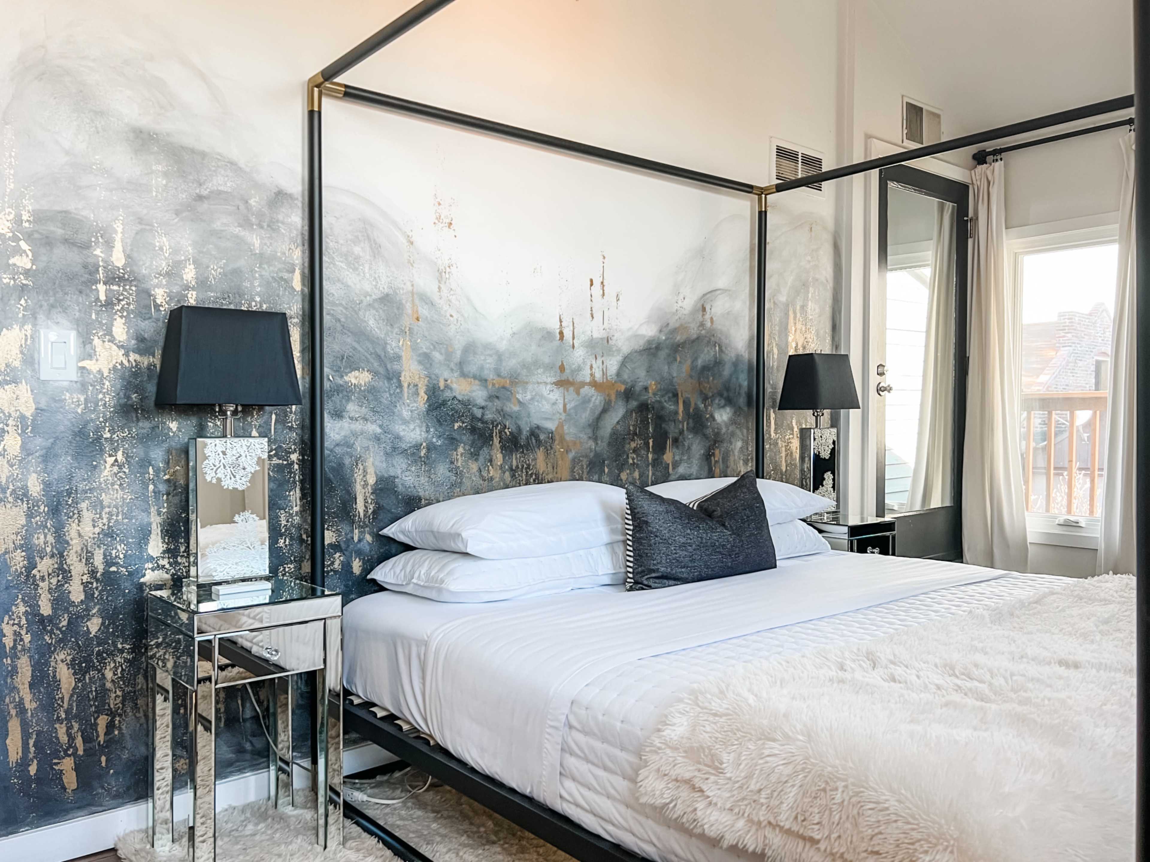The image shows a stylish bedroom featuring a four-poster bed with white linens, mirrored bedside tables, and a decorative wall with a gold and gray abstract design.