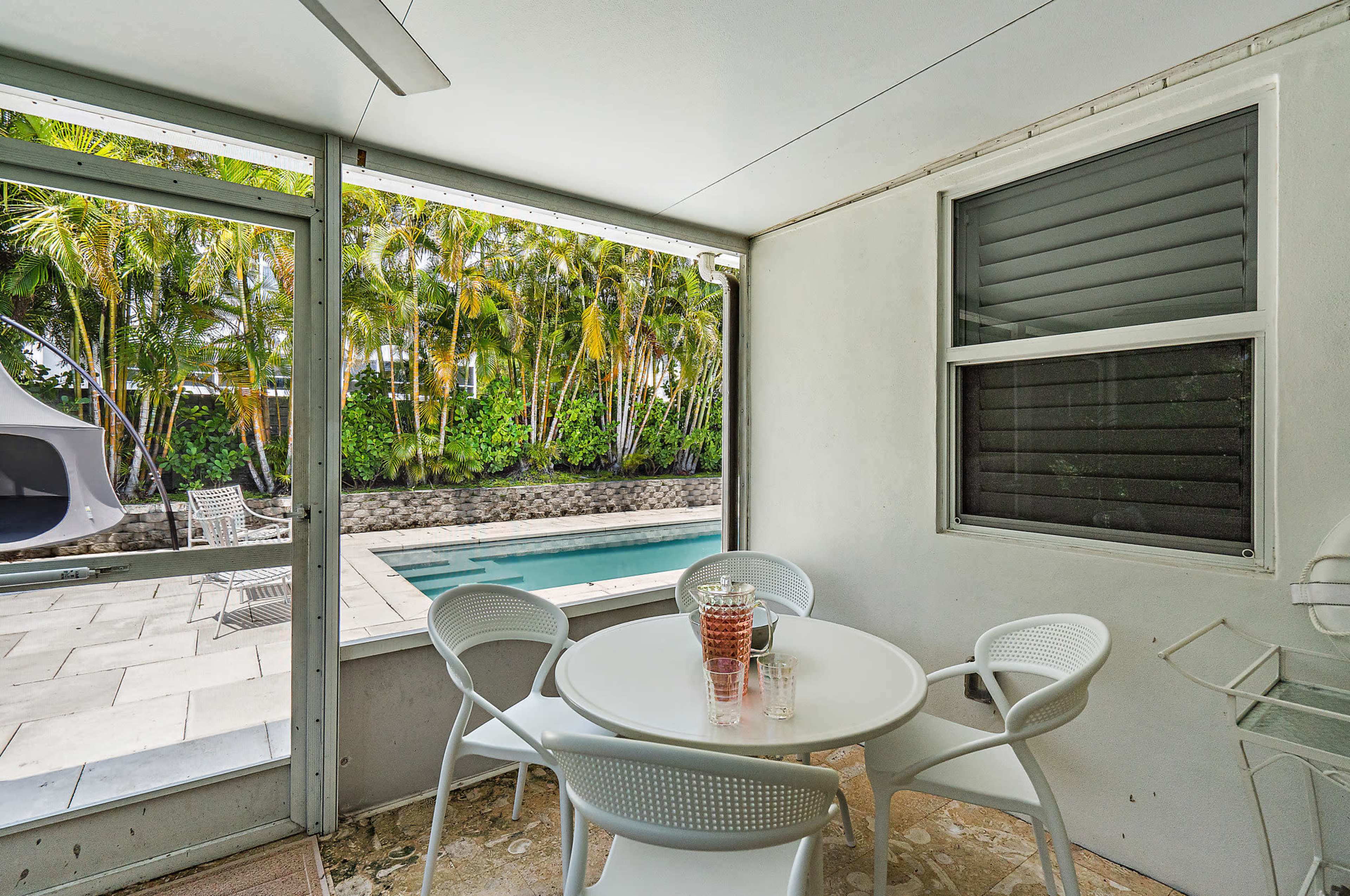 A glass-enclosed patio features a round table with four chairs, overlooking a swimming pool surrounded by lush greenery.