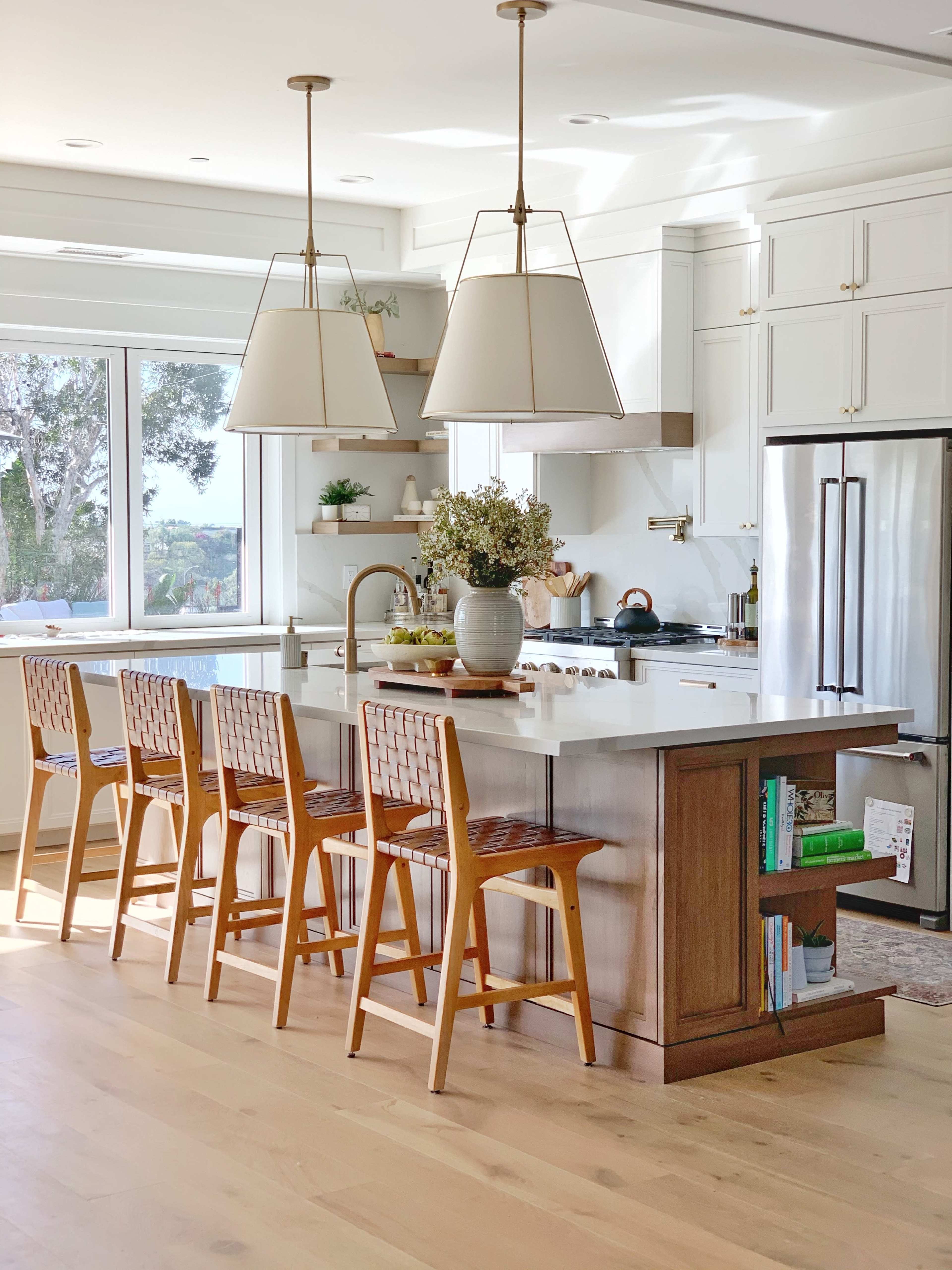 A modern kitchen features a central island with bar stools, pendant lights, and large windows allowing natural light to enter.