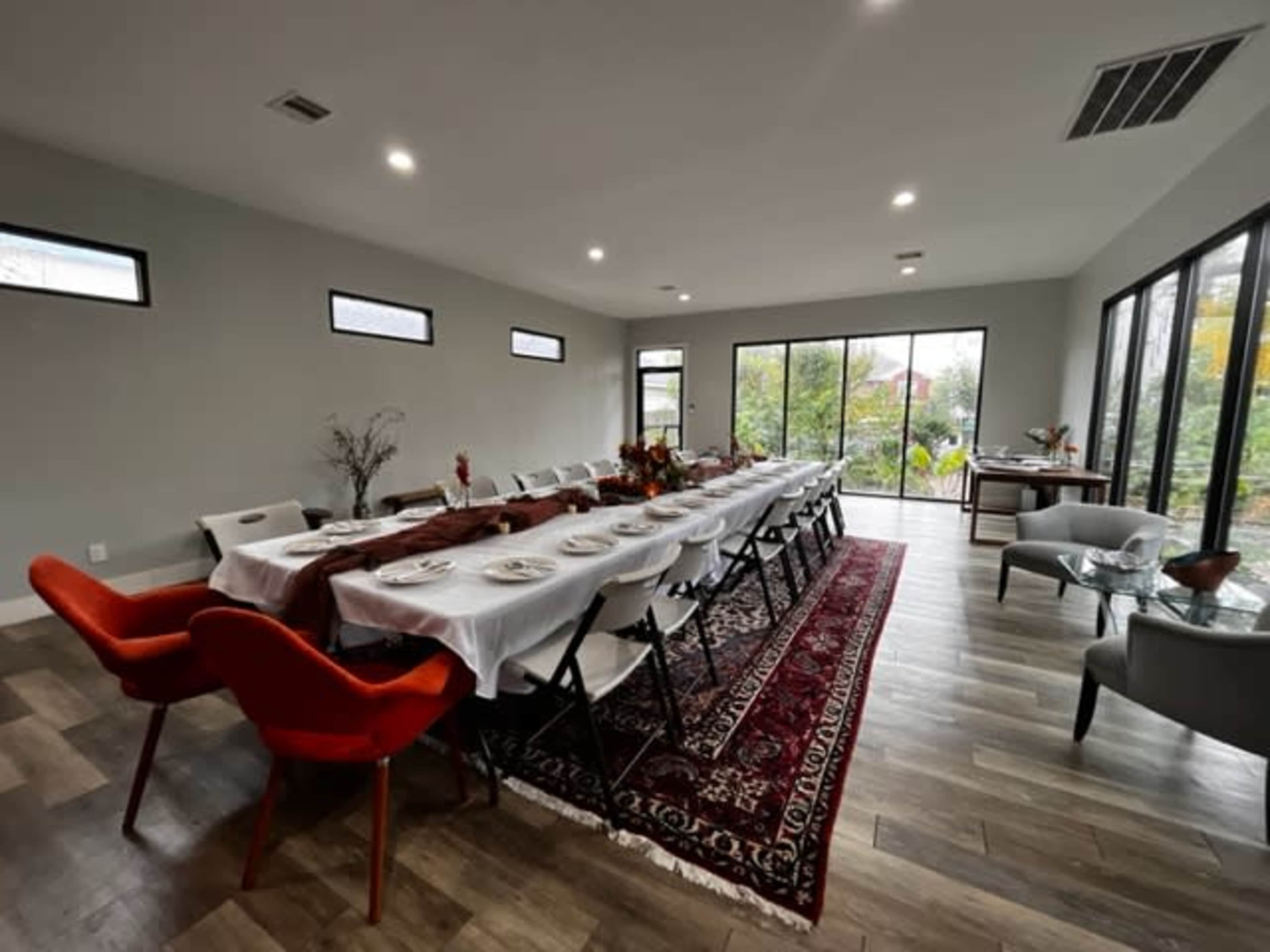 A long dining table with white linens is set for a meal in a spacious room featuring large windows and a view of greenery.
