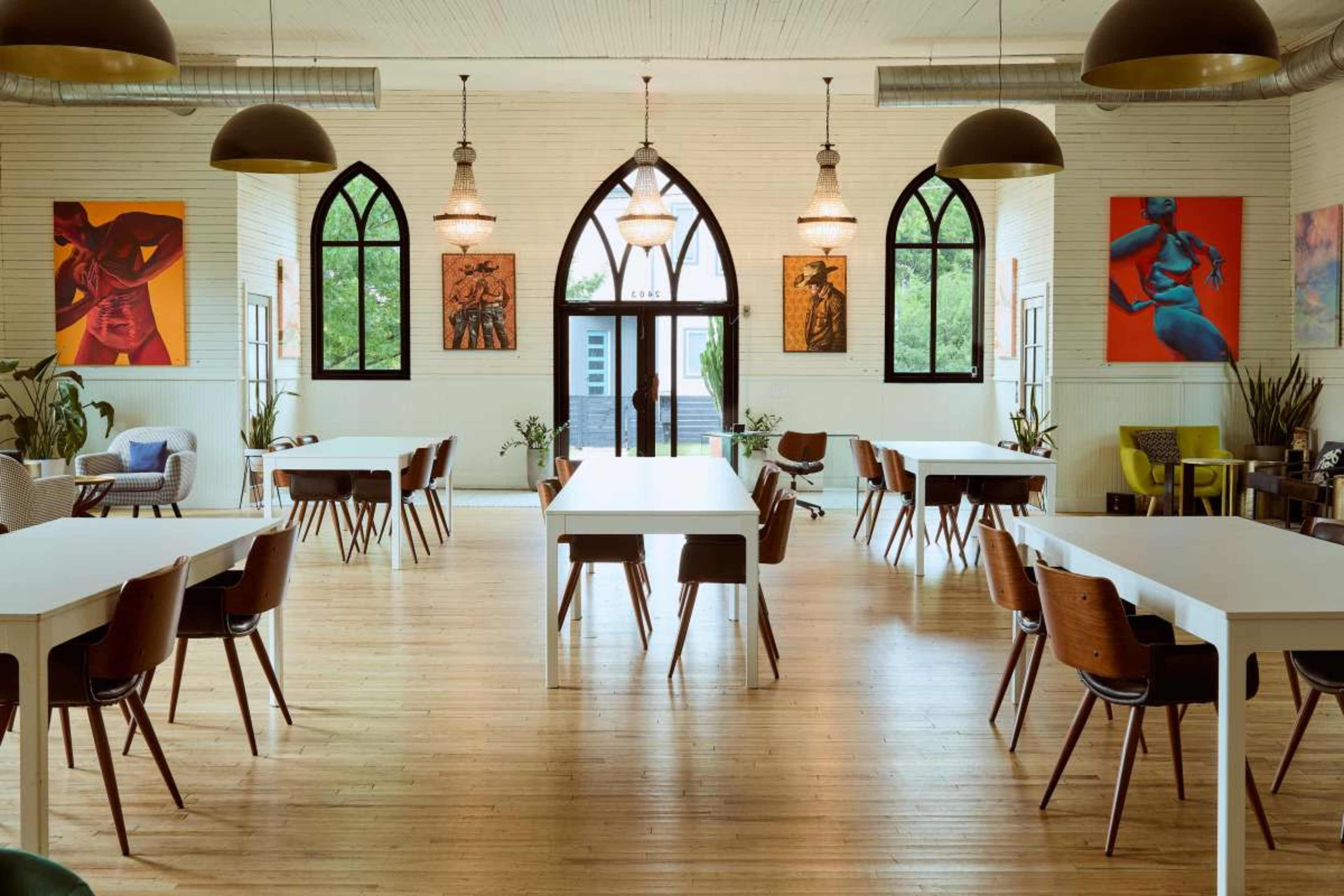 The image depicts a spacious, bright room with wooden flooring, featuring white tables and chairs arranged in rows, along with large arched windows and colorful artwork on the walls.