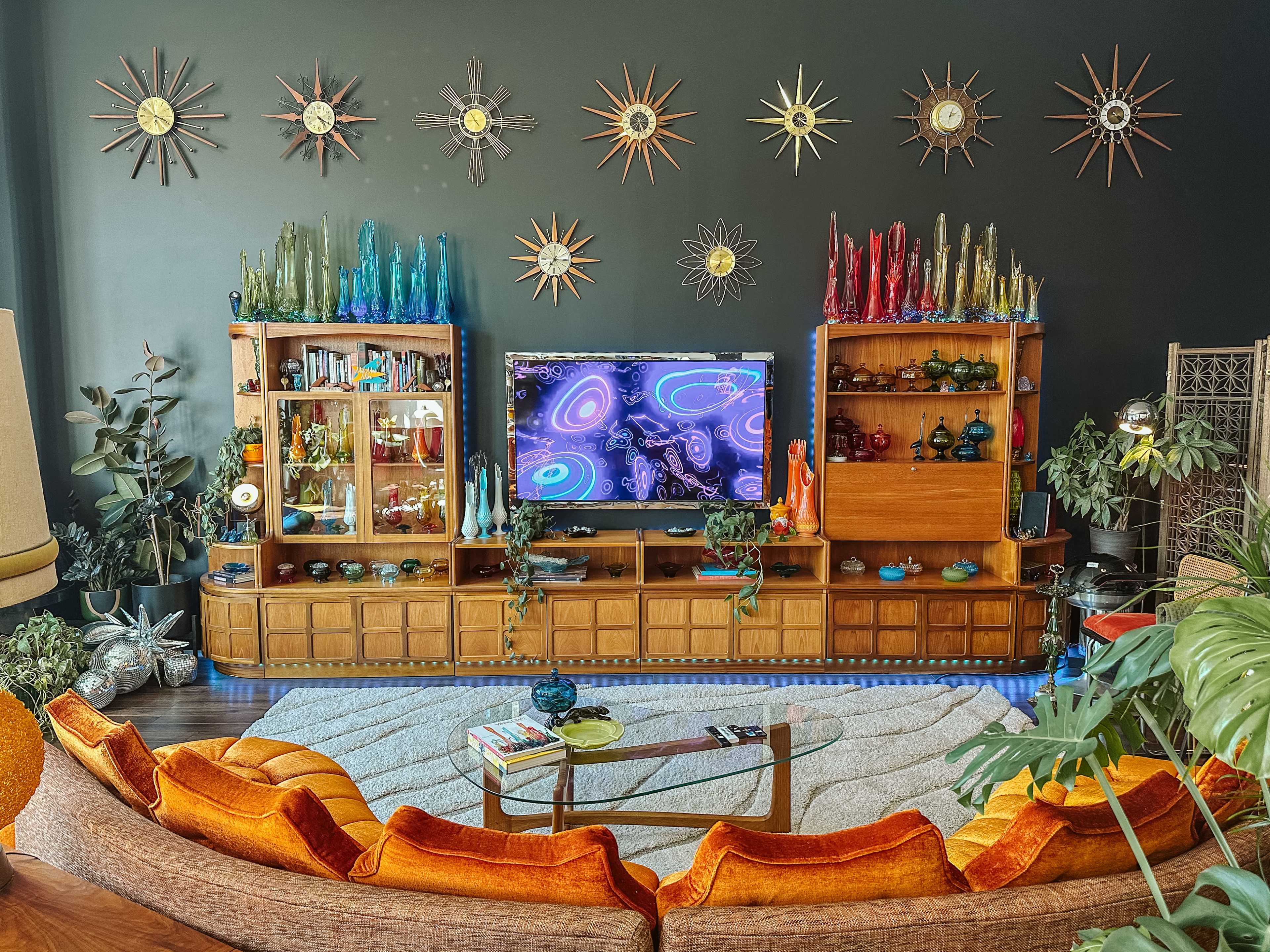 A mid-century modern living room features a curved orange sofa in front of a large wall-mounted television, flanked by wooden display cabinets adorned with colorful glassware and starburst wall decorations.