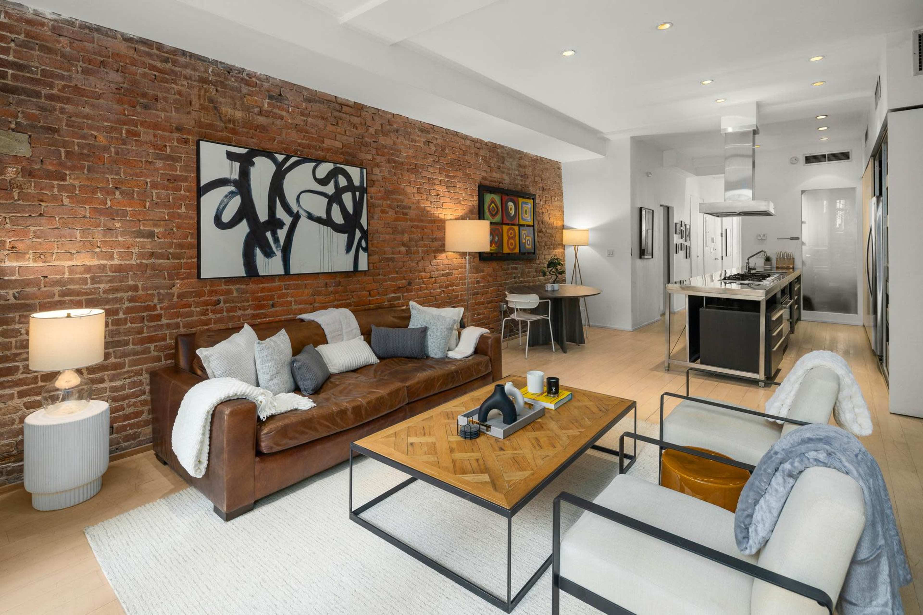 A modern living room with exposed brick walls, a leather sofa, a wooden coffee table, and a kitchen in the background.