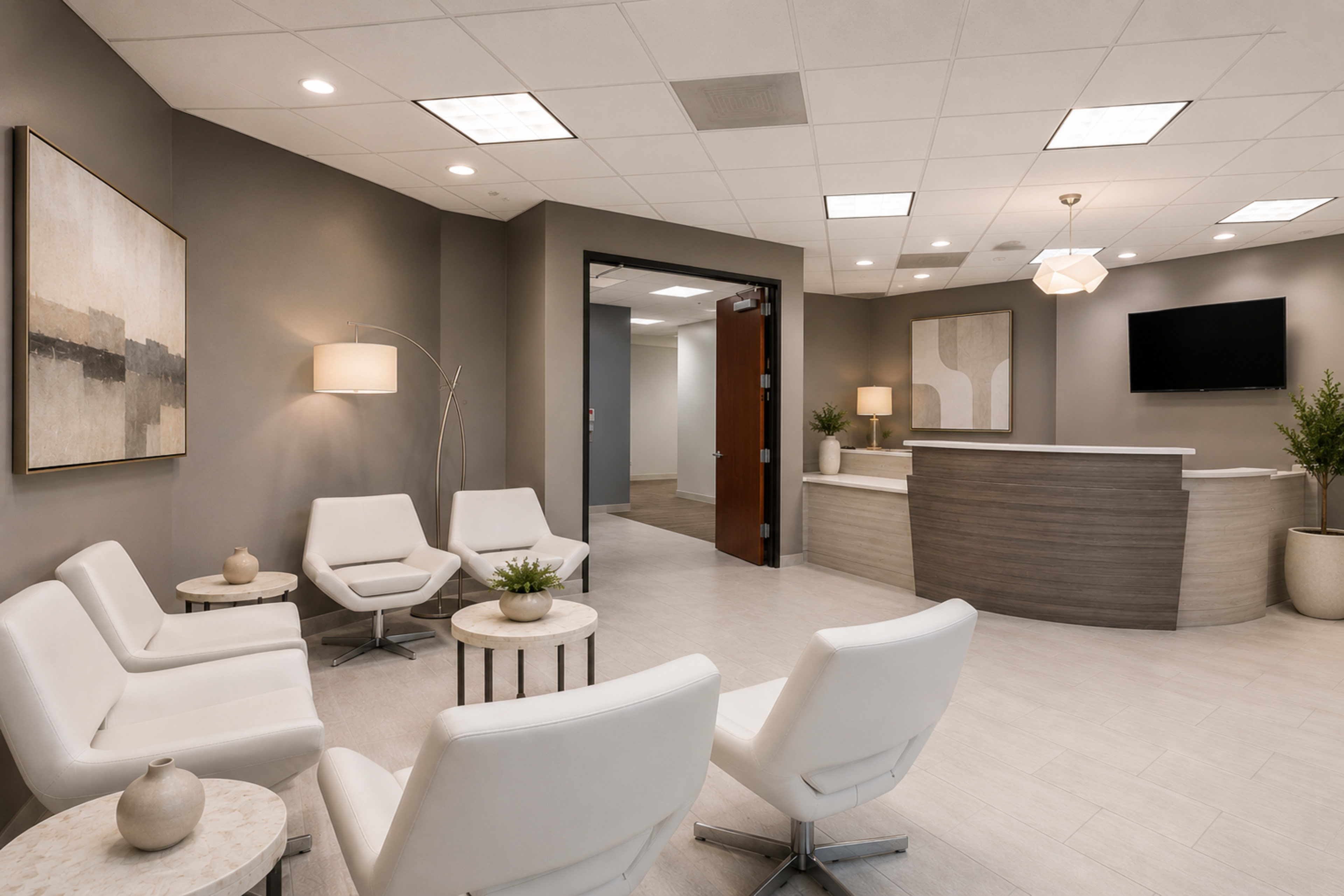 The image shows a modern waiting area with white chairs, small tables, a reception desk, and muted wall decor.