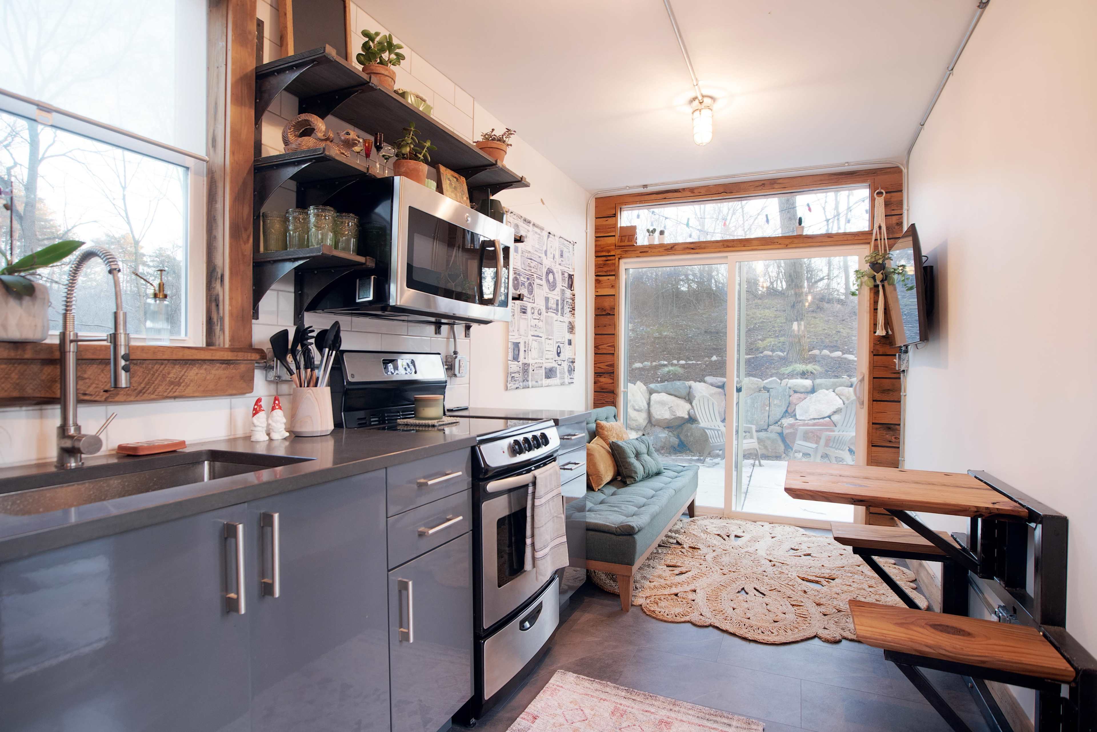 A compact kitchen with gray cabinets, stainless steel appliances, a small dining table, and a cozy seating area next to a large window.