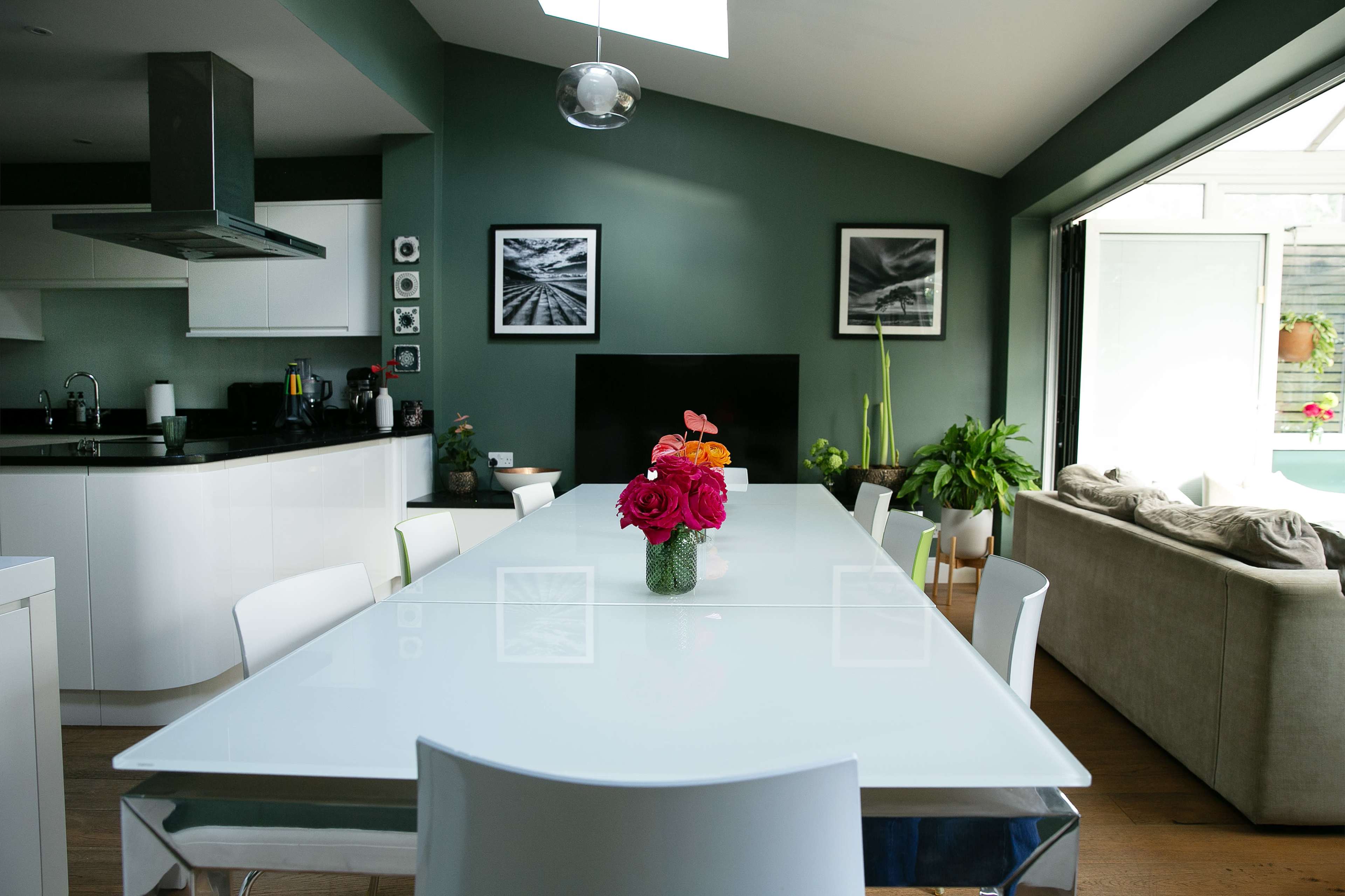 A modern dining area features a long white table with a floral centerpiece, surrounded by white chairs, and is illuminated by natural light coming from large windows.