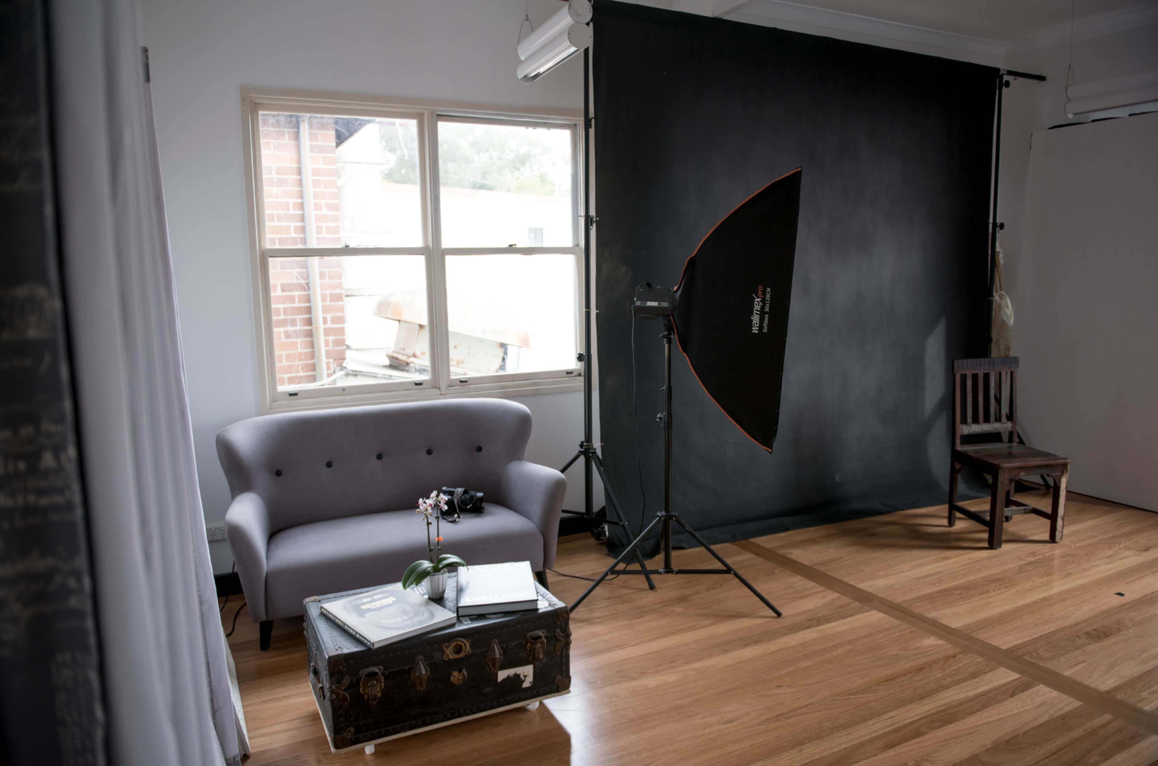 The image shows a photography studio with a gray sofa, a black backdrop, a softbox light setup, a chair, and a window allowing natural light to enter.