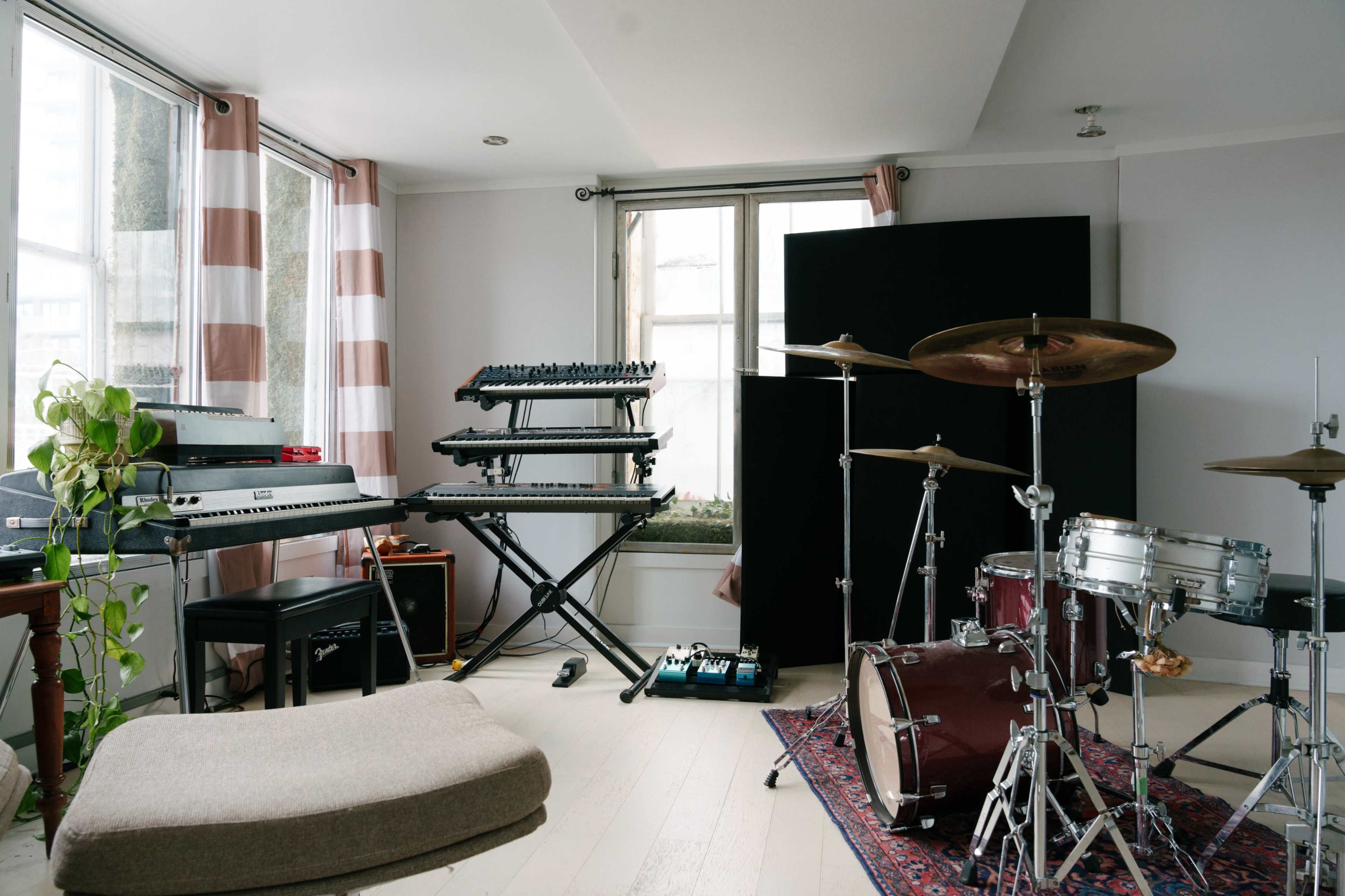 A music studio features a keyboard, synthesizer, and drum set arranged around natural light from large windows.
